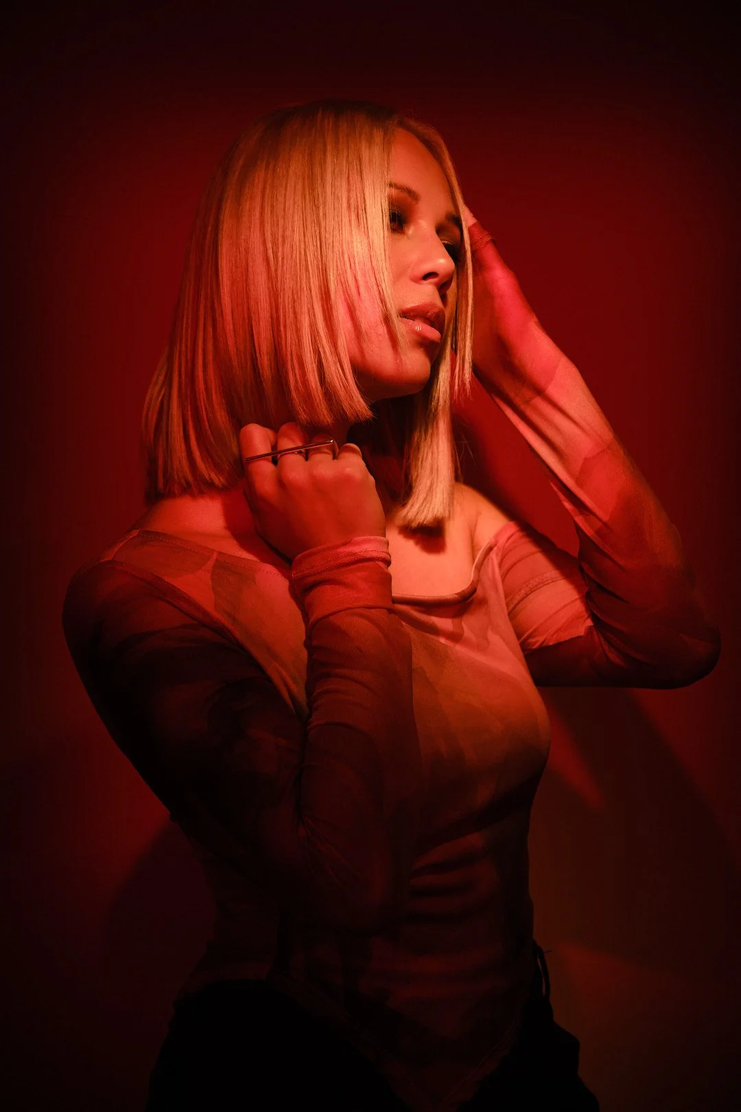 A woman with blonde hair styled in a bob pose under red lighting, wearing a sheer, patterned long-sleeve top, with one hand touching her hair and the other resting near her chin.