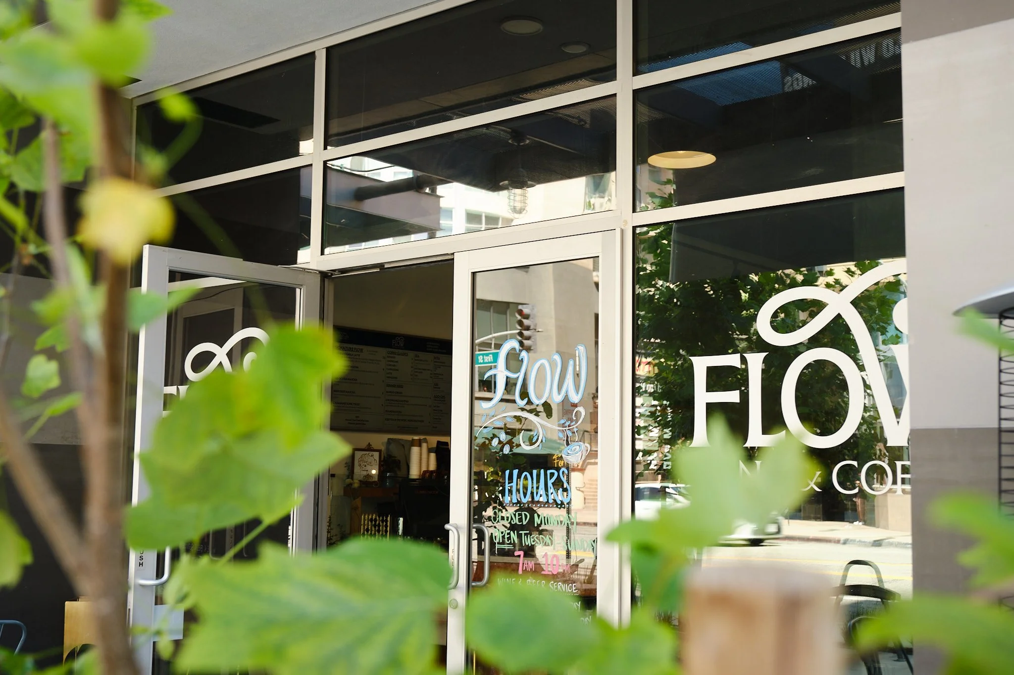 View of a storefront with large glass windows and door, with colorful handwritten signs indicating business hours and offerings. Green leaves partially obscure the view.