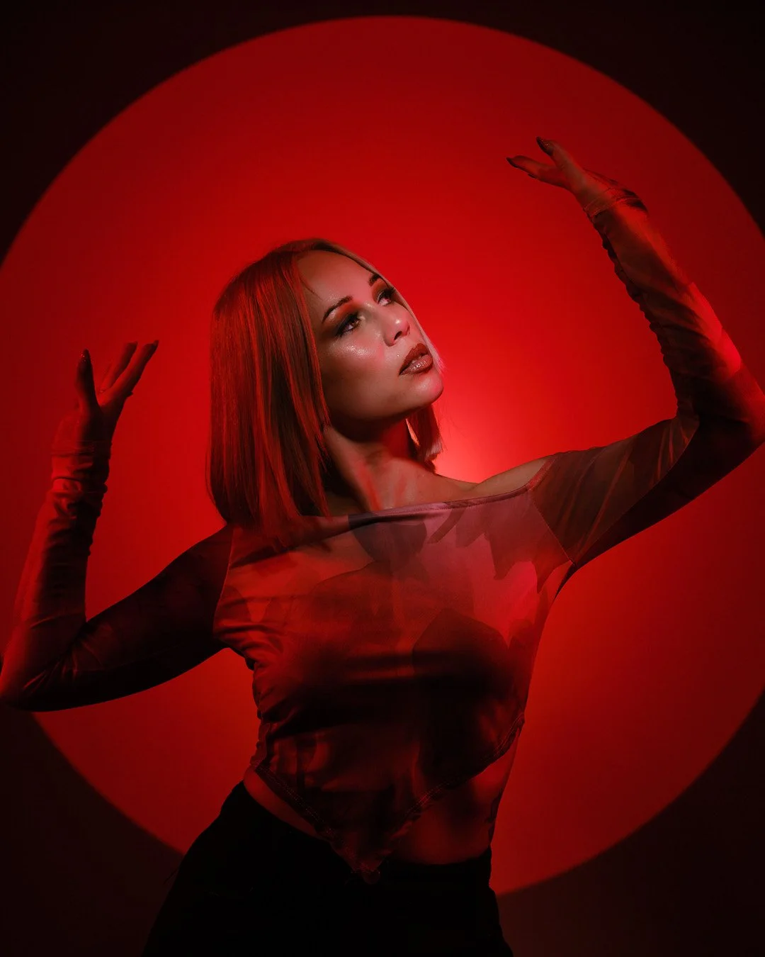 A woman with red hair posing with her arms raised against a red circular background, wearing a sheer top and long gloves.