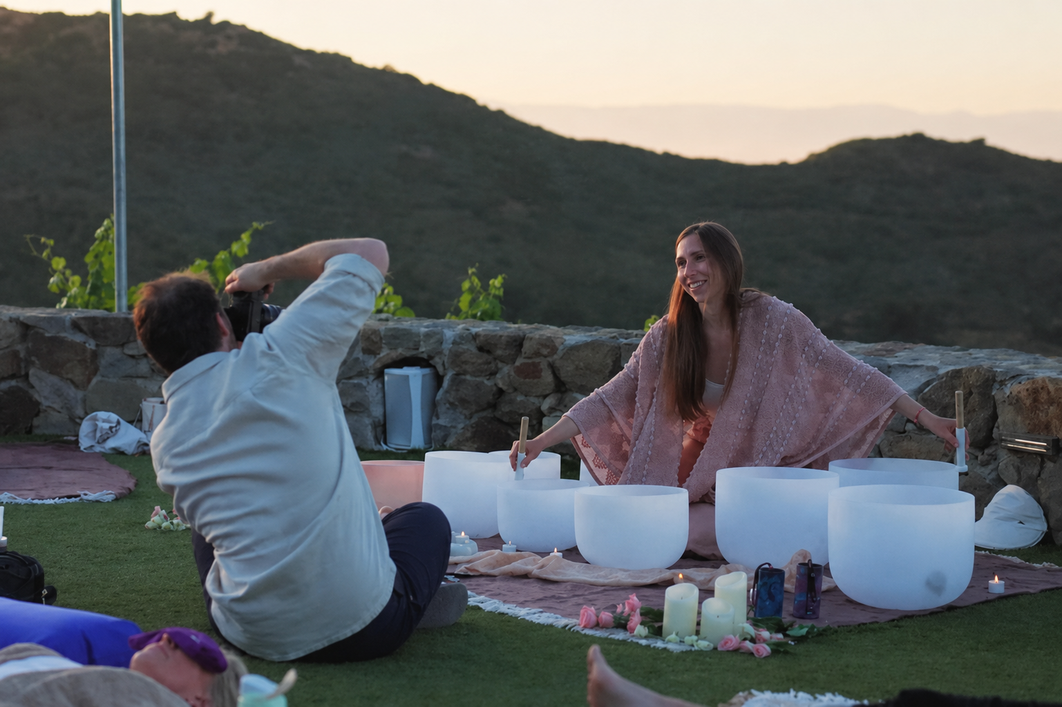 A woman sitting on the ground playing singing bowls at an outdoor sunset music session, while a man with a camera photographs her, with a scenic hilly background.