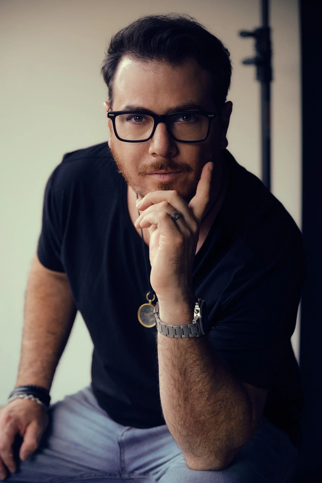 A young man with dark hair, beard, and glasses poses with his hand on his chin, wearing a black T-shirt, a silver wristwatch, and a bracelet, in front of a neutral background.