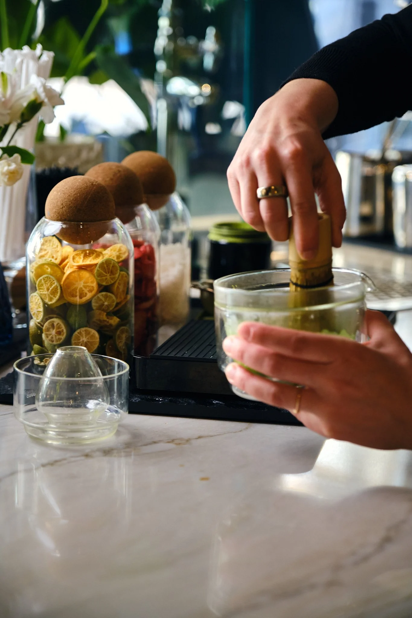 A person using a wooden muddler to crush ingredients in a glass bowl, with several jars of dried citrus slices, dried flowers, and herbs on a marble countertop.