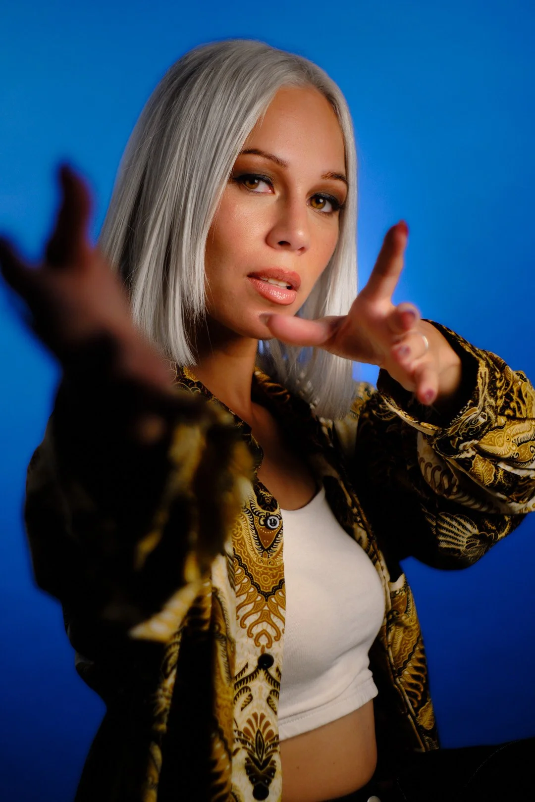 A woman with platinum blonde hair and makeup posing against a blue background, wearing a patterned jacket and a white crop top, with her hands reaching toward the camera.