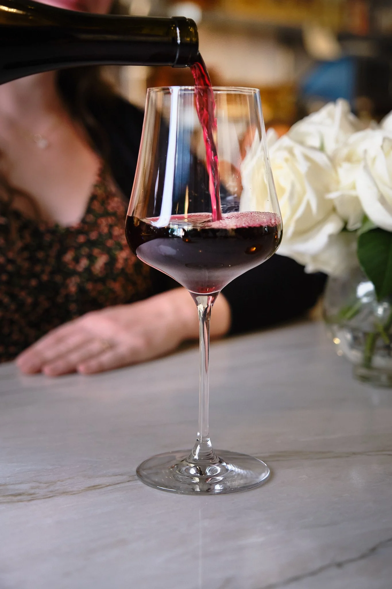 Red wine being poured into a stemmed wine glass on a light-colored surface. A blurred person and a bouquet of white flowers are in the background.