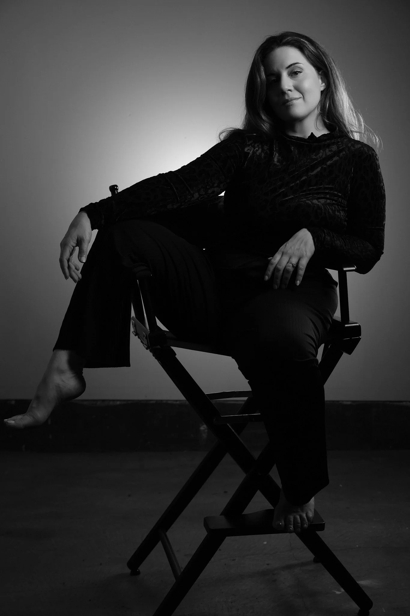 A black and white photo of a woman sitting on a director's chair, smiling softly, with one leg bent and foot resting on the ground, and the other extended out in front. She is wearing a long-sleeved, velvet, patterned top.