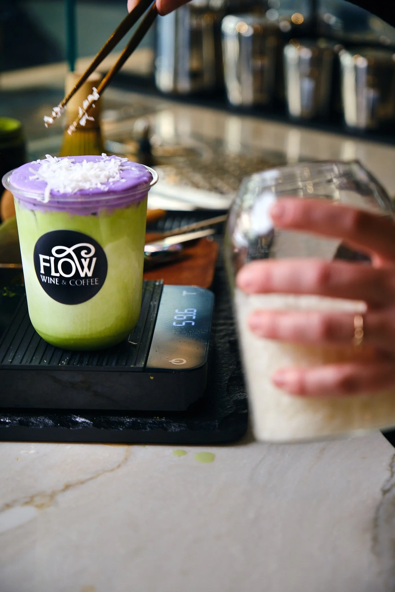 A hand holding a glass of a light-colored beverage in front of a blurred background with a colorful drink on a scale.