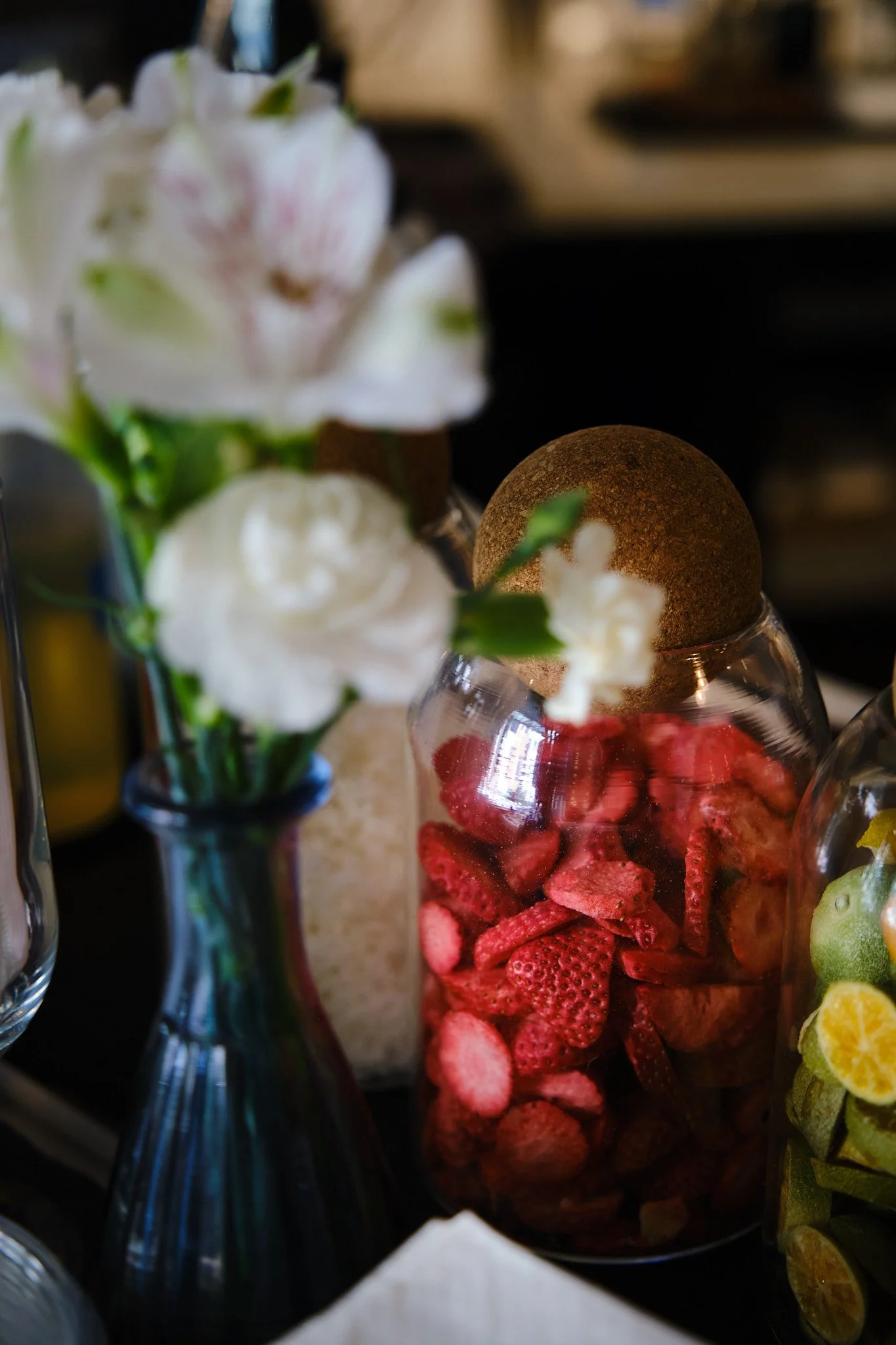 A glass jar filled with sliced strawberries and whole strawberries, with white flowers and a brown egg on top. Tall glass vases with green plants are visible.