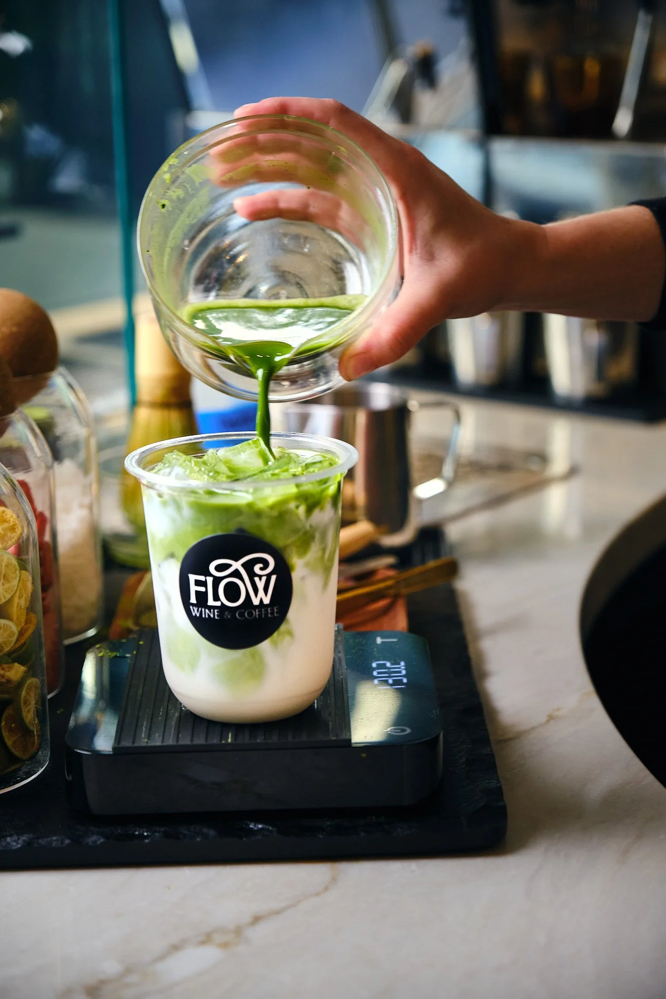 A person pours green matcha latte into a clear cup with ice from a glass container at a cafe named Flow Wine & Coffee, with a digital scale underneath the cup.