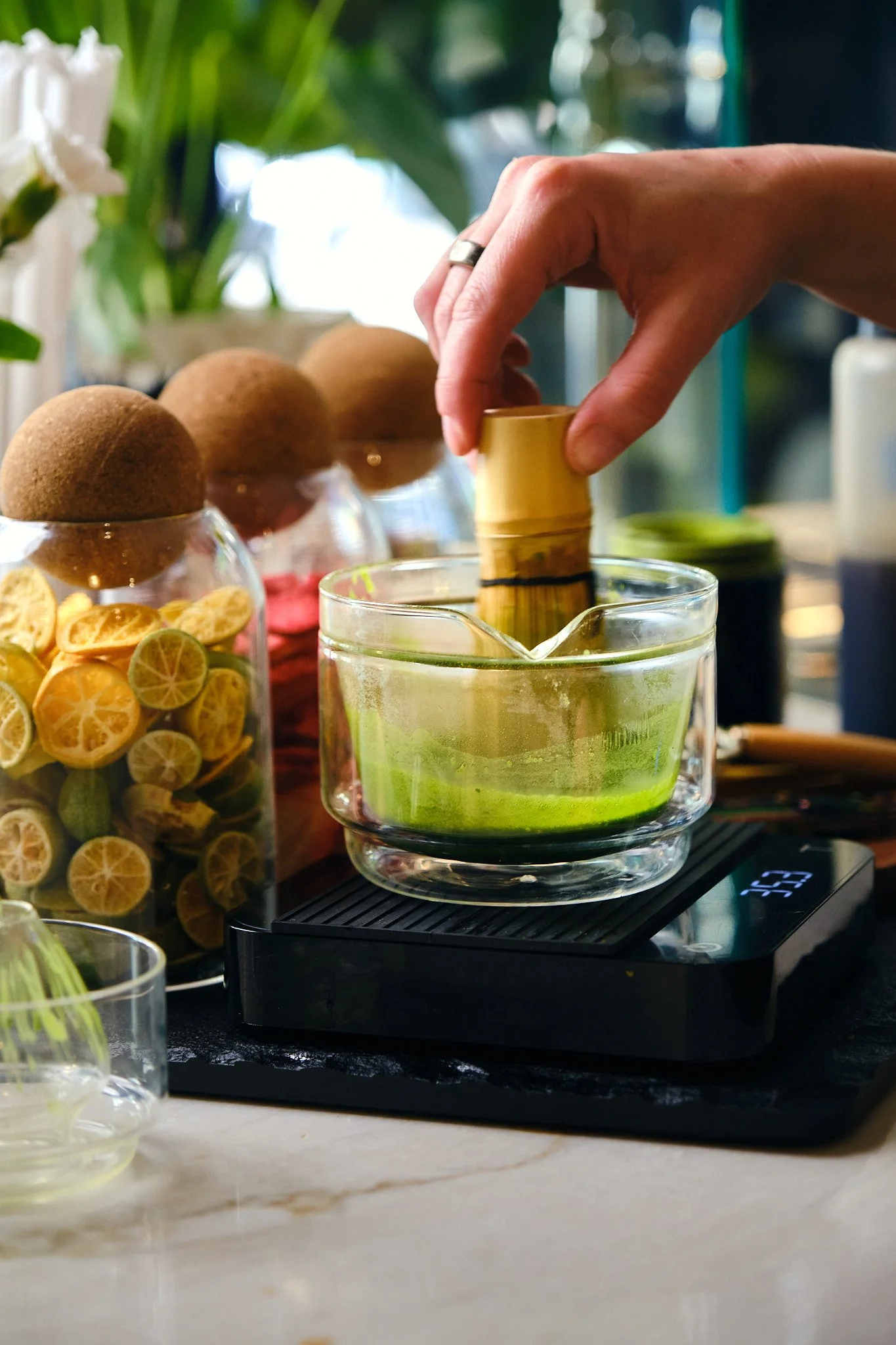 A person is using a bamboo whisk to whisk matcha green tea in a glass bowl, placed on a digital scale; there are dried citrus slices in a jar and kiwifruit in the background.