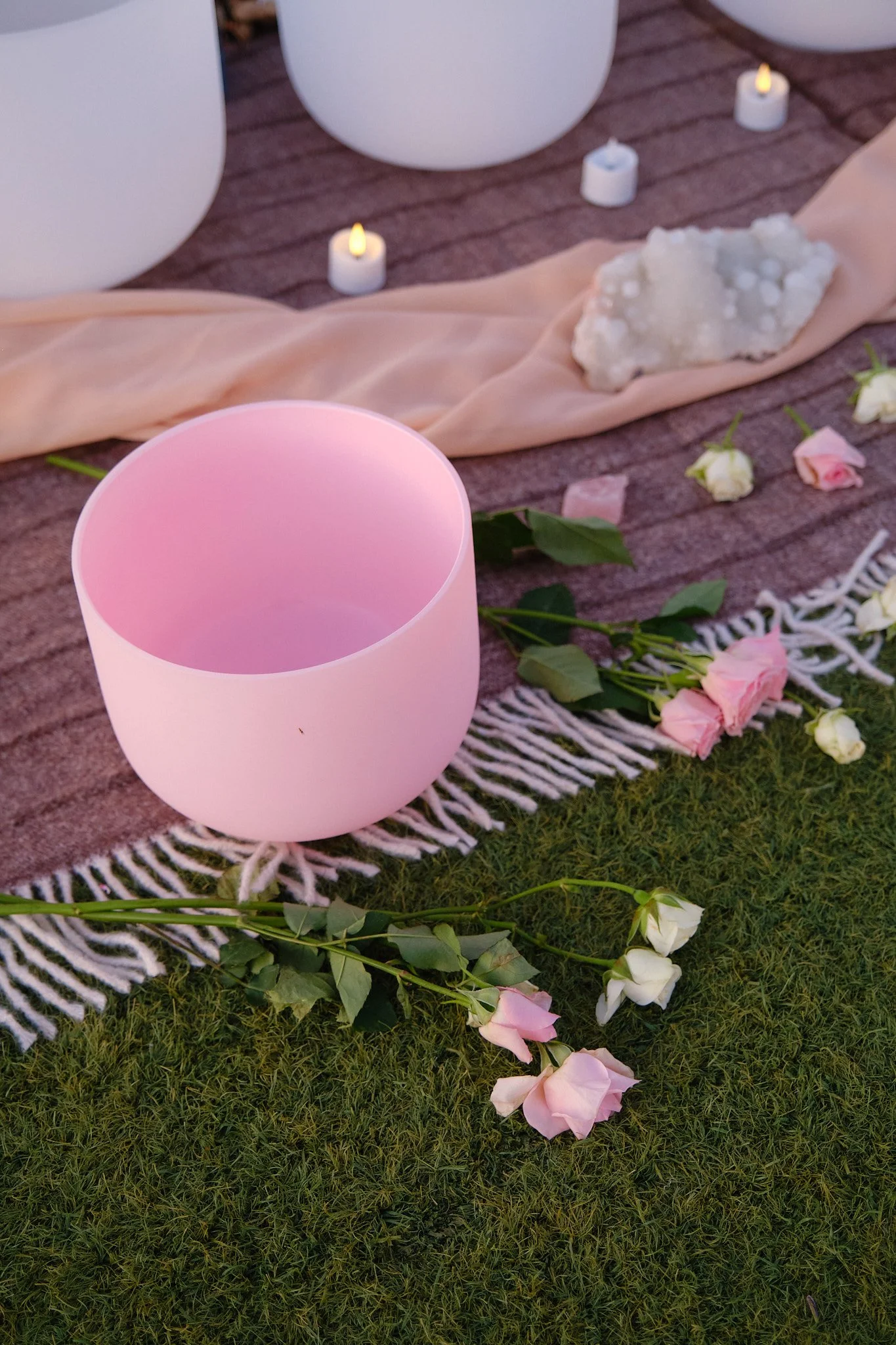 Decorative setup on a grassy surface featuring a pink candle holder, scattered pink and white roses, candles, white vases, and a beige fabric runner.