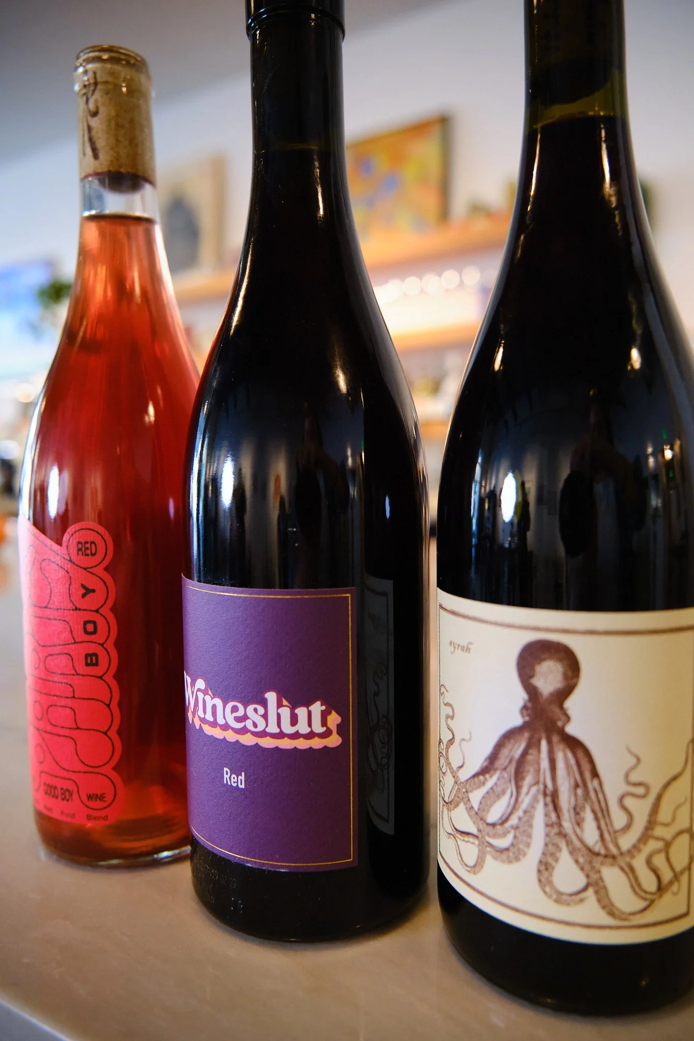 Close-up view of three bottles of wine or beverage on a table. The left bottle has a red label, the middle bottle has a purple label with the text 'Wineslut' and 'Red', and the right bottle has a lighter label with a skeleton illustration.