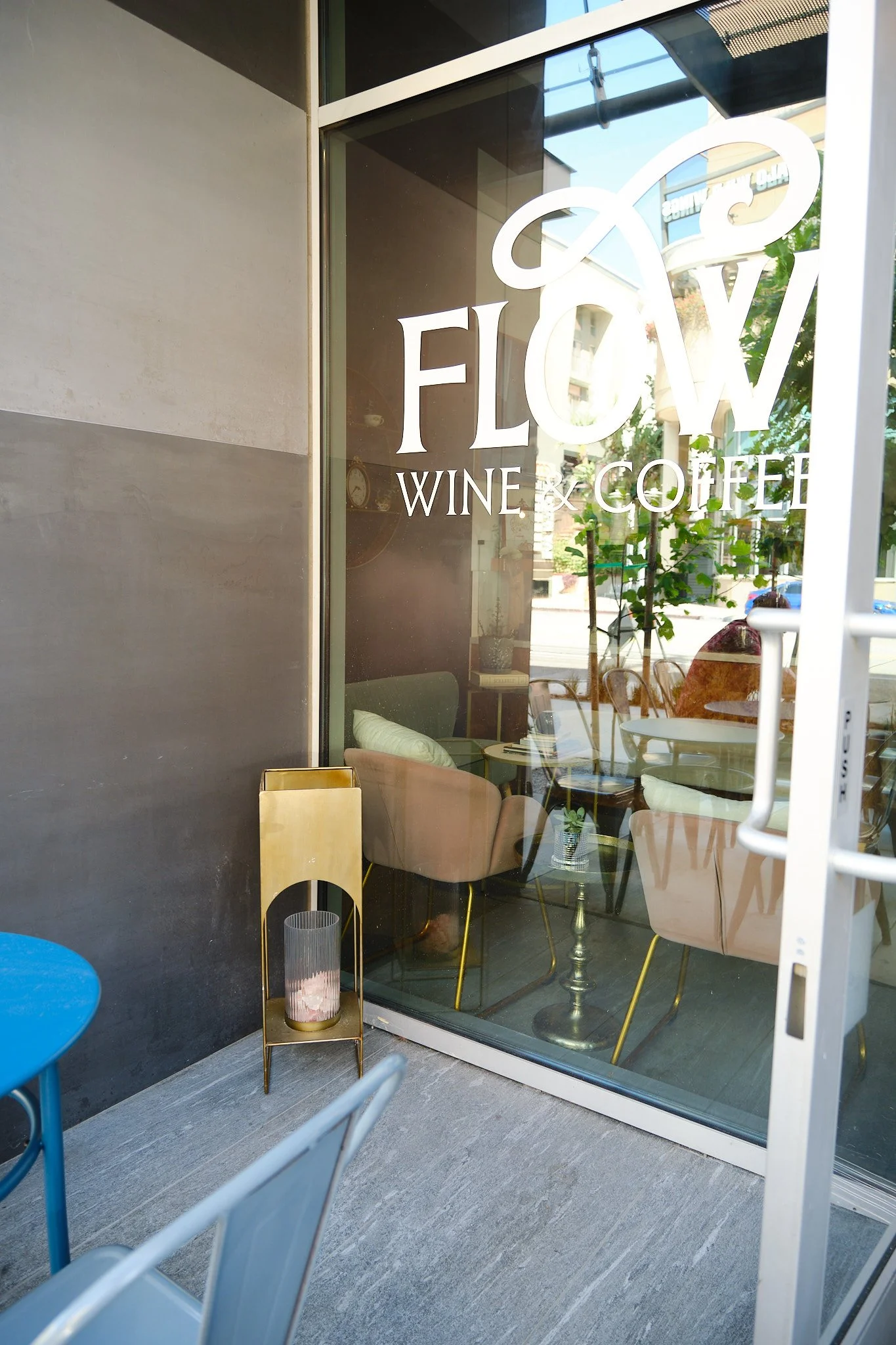 View of a coffee shop interior seen through a large glass window with the sign 'FLOY WINE & COFFEE.' Inside, there are chairs, tables, and plants, creating a cozy atmosphere. Outside, part of a blue table is visible, and a modern lamp is placed on a 