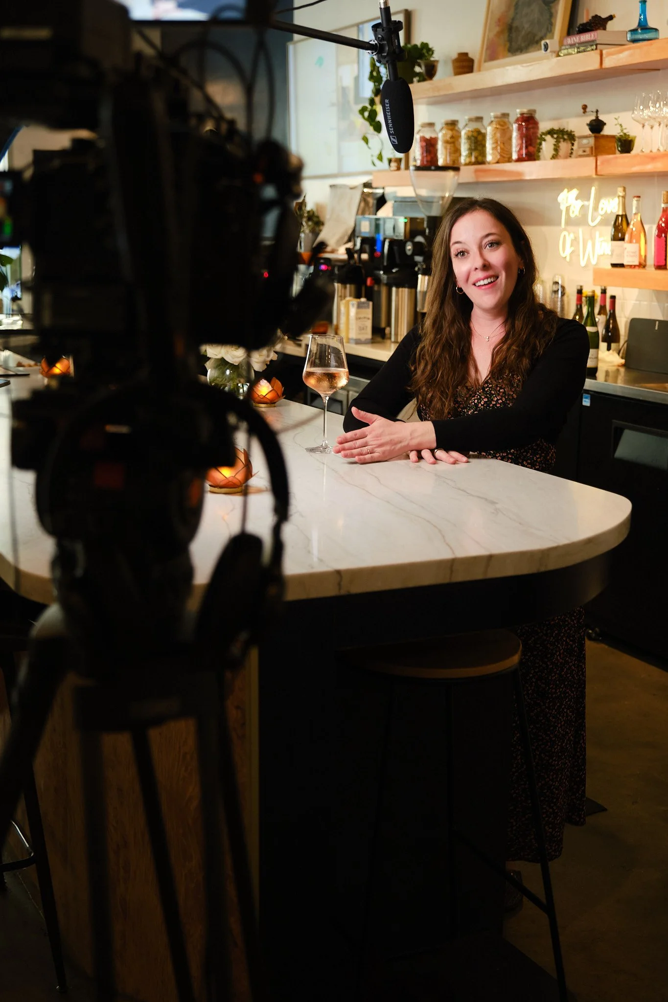 A woman sitting at a bar counter with a glass of rosé wine, being filmed or photographed in a cozy, well-lit cafe or bar with a homey decor.