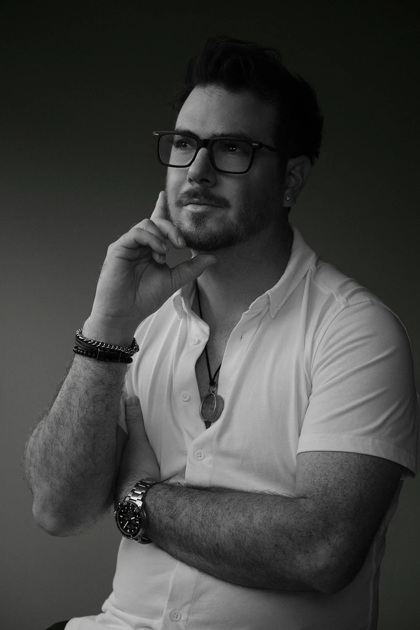 A man with dark hair, glasses, and a beard, wearing a white button-up shirt, is sitting with his right arm crossed over his chest and his left hand resting on his chin. He is wearing a large watch on his left wrist and a bracelet on his right wrist. The background is plain and dark.