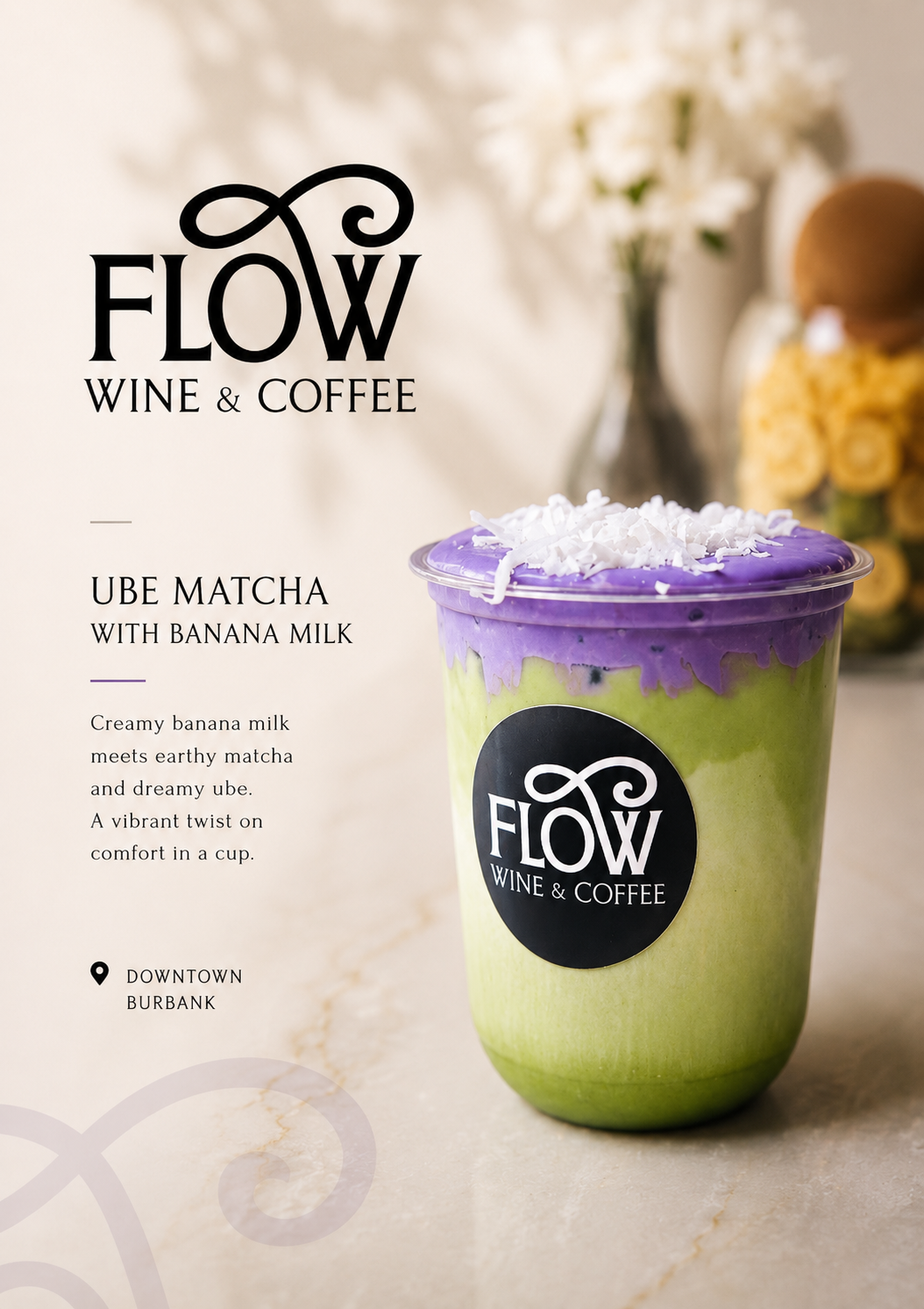 Purple and green matcha drink topped with coconut flakes, branded with a Flow Wine & Coffee logo, with blurred white flowers and a jar of yellow flowers in the background.