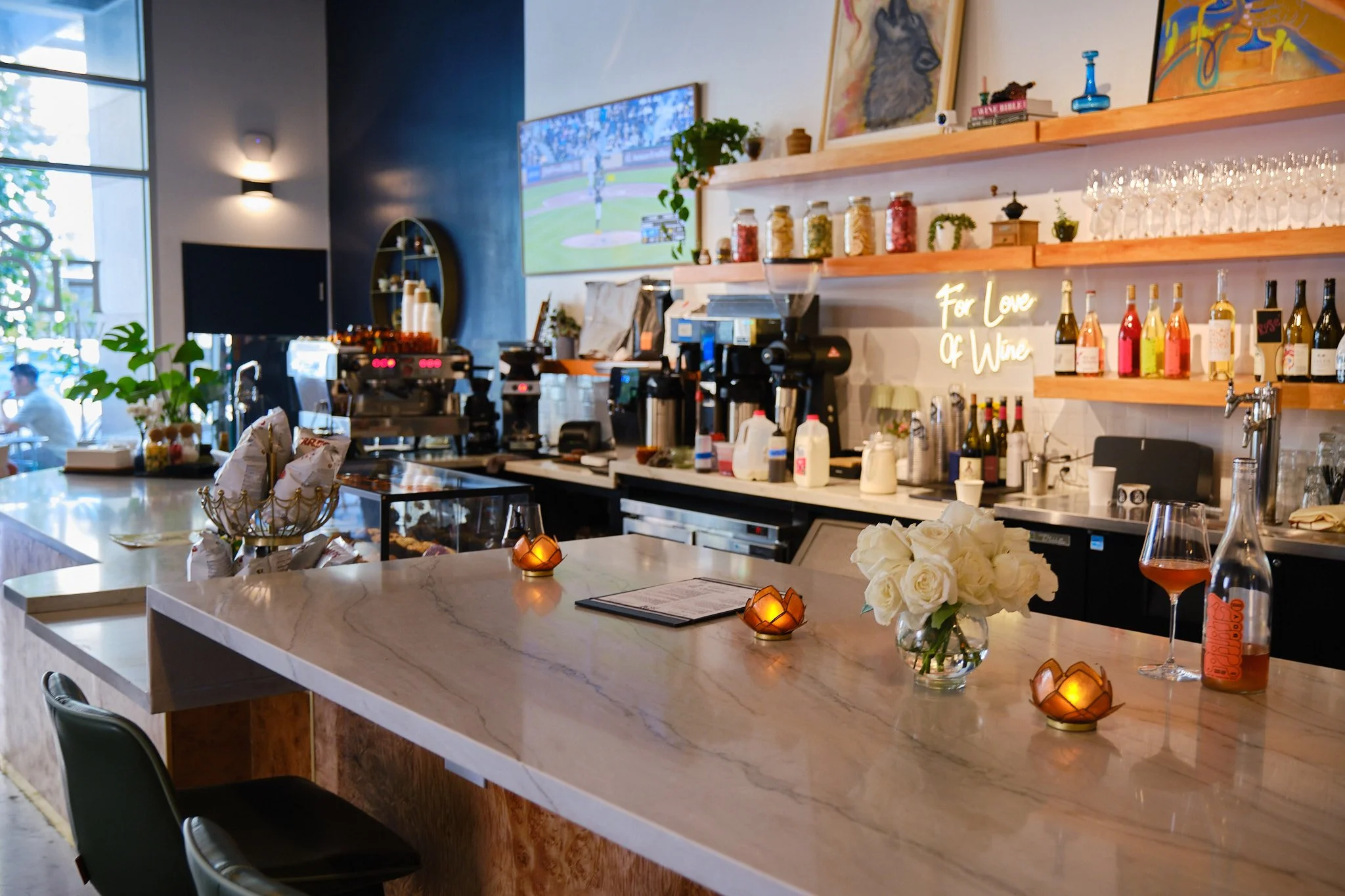 A modern cafe or bar interior with a white marble counter, small decorative candles, bottles of wine or spirits, a vase of white roses, and a menu on the counter. In the background, there are shelves with glassware and jars, a neon sign that reads 'F