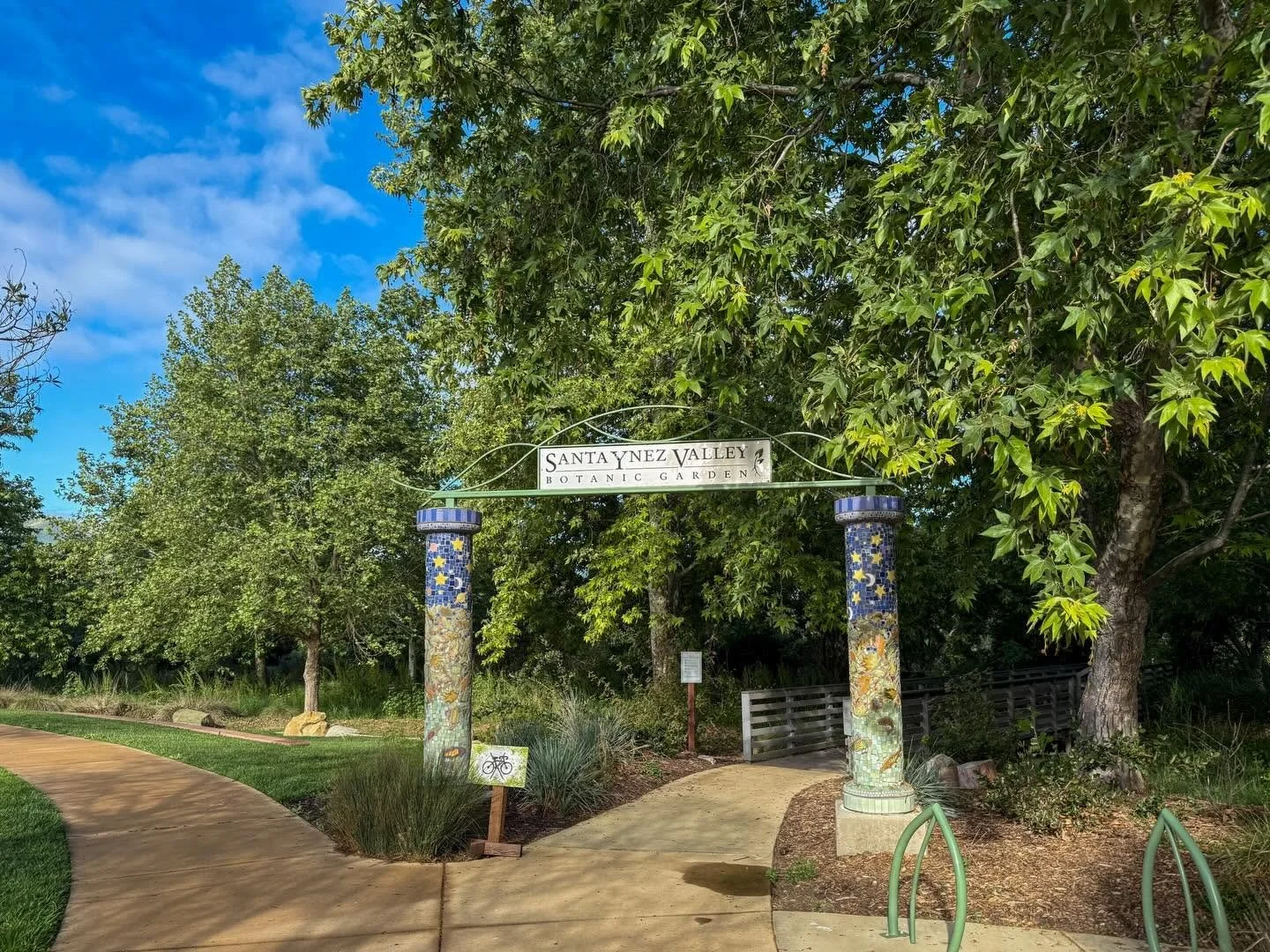 Entrance archway at Santa Ynez Valley Botanic Garden, decorated with colorful mosaic tiles on two columns, with a pathway leading into a garden area surrounded by trees and greenery under a partly cloudy sky.