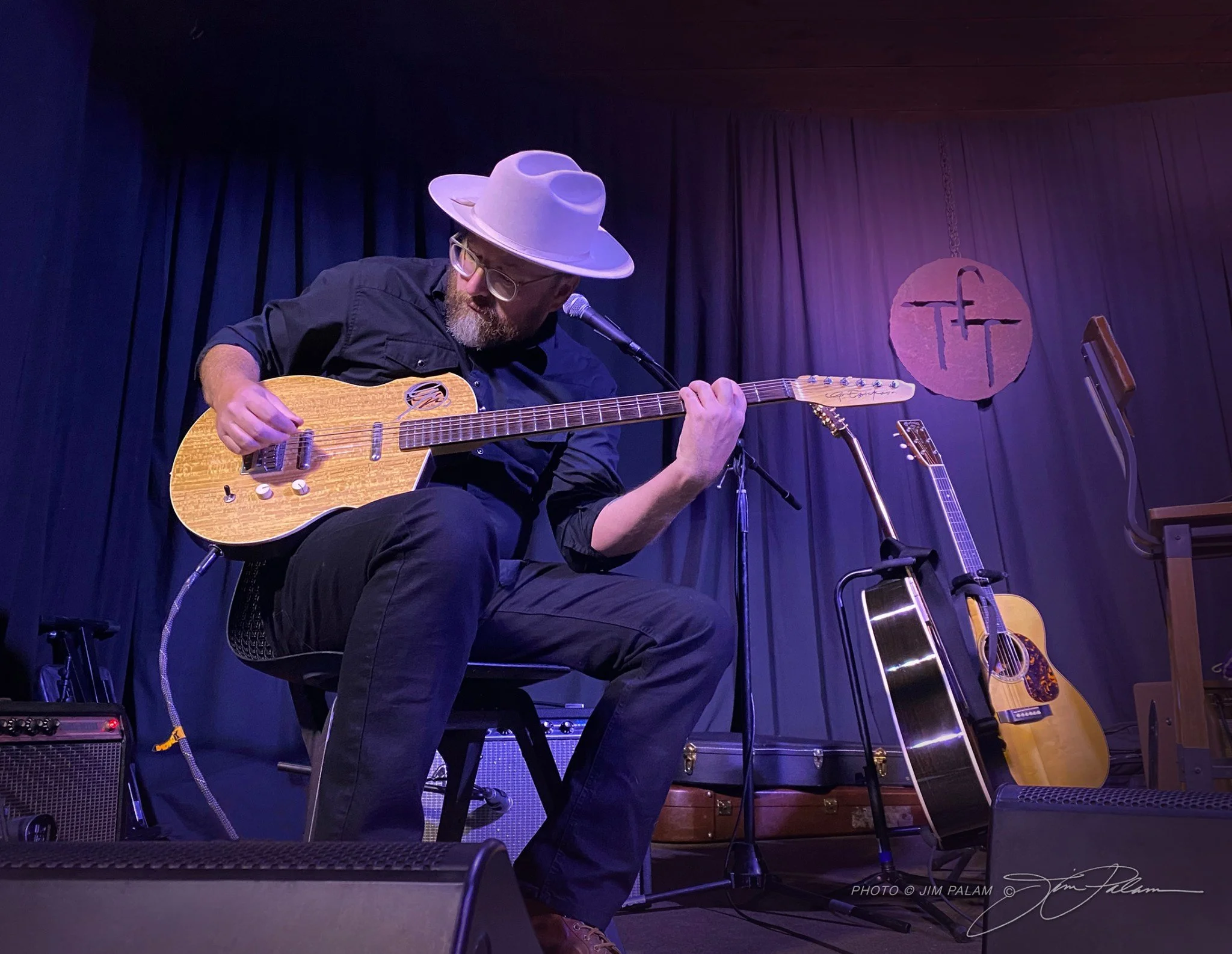 A man wearing glasses and a light-colored hat playing an acoustic guitar on stage with a microphone in front of him, set against a dark blue and purple background with stage curtains and guitars visible.