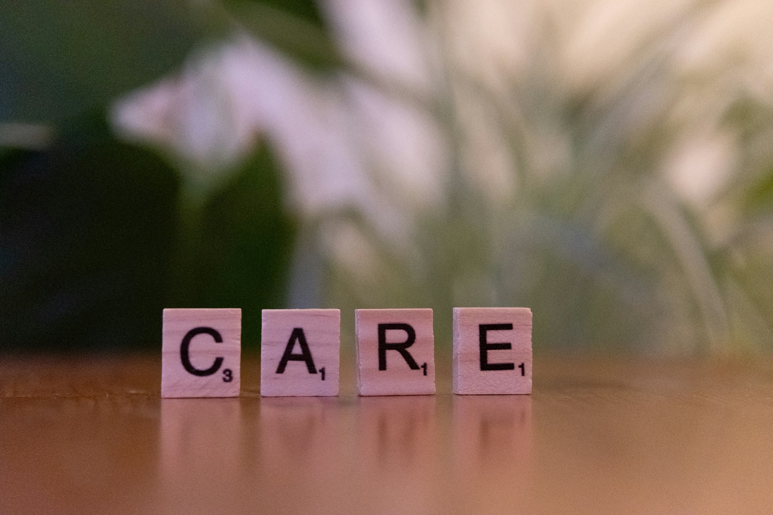Scrabble tiles spelling the word "CARE" with a blurred background of green and white, and part of a word starting with 'C' in the upper left corner.