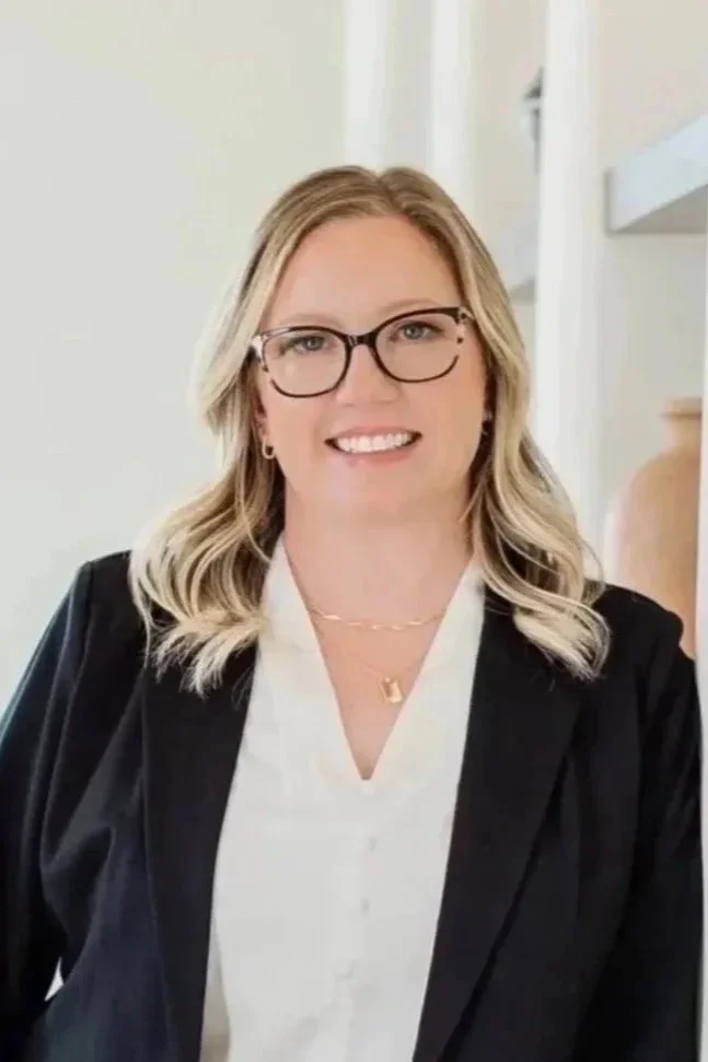 A woman with blonde hair, wearing glasses, a white blouse, and a black blazer, smiling and looking at the camera.