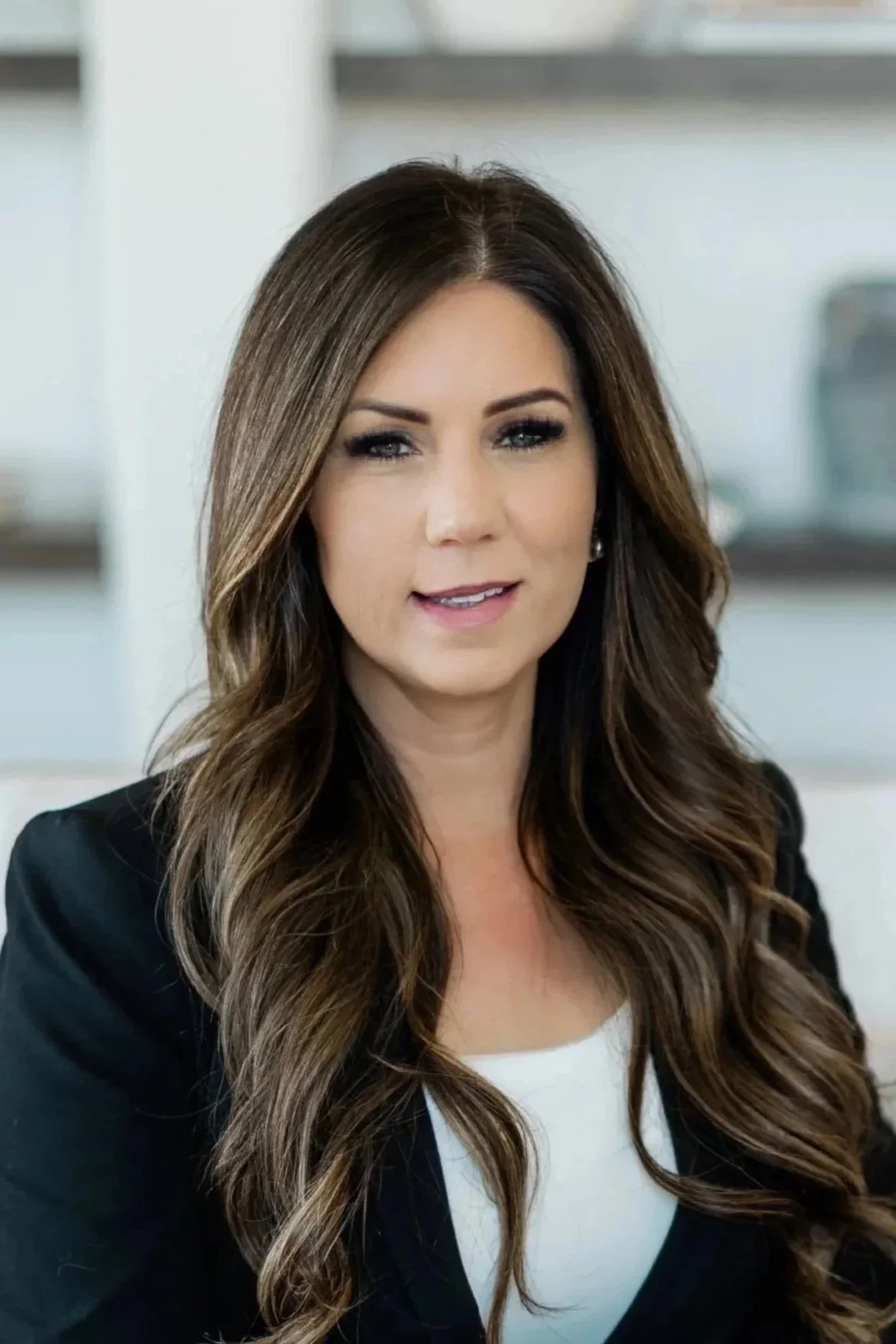 A professional woman with long, wavy brown hair, wearing a black blazer and white top, smiling indoors.