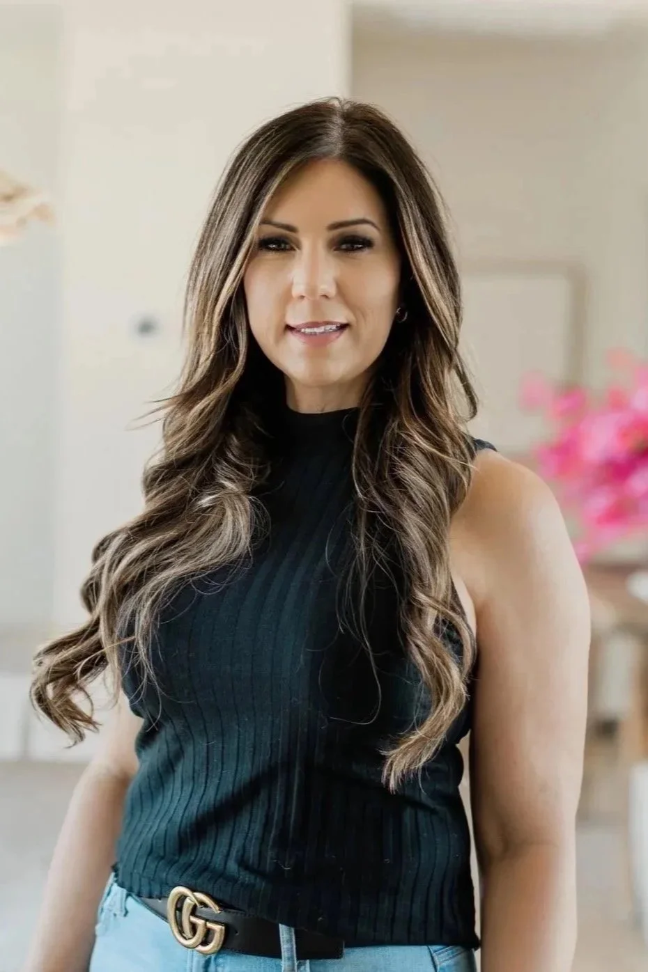 A woman with long wavy brown hair, wearing a black sleeveless top and light blue jeans with a black belt, standing indoors near pink flowers.