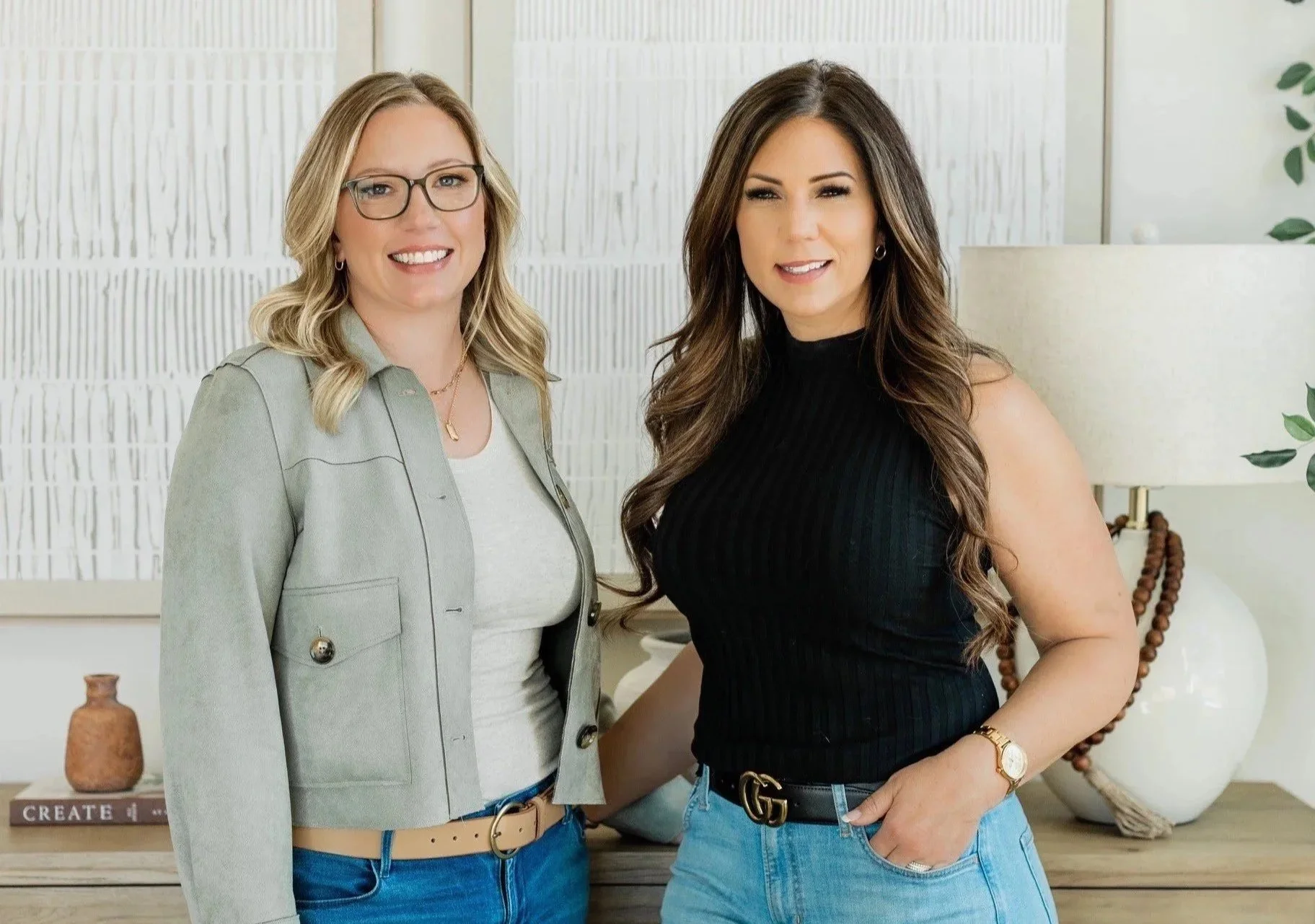 Two women standing next to each other indoors with decorative objects and a white background. The woman on the left has blonde hair, glasses, and is wearing a light green blazer over a beige top. The woman on the right has long brown hair and is wearing a black sleeveless top and blue jeans with a Gucci belt.