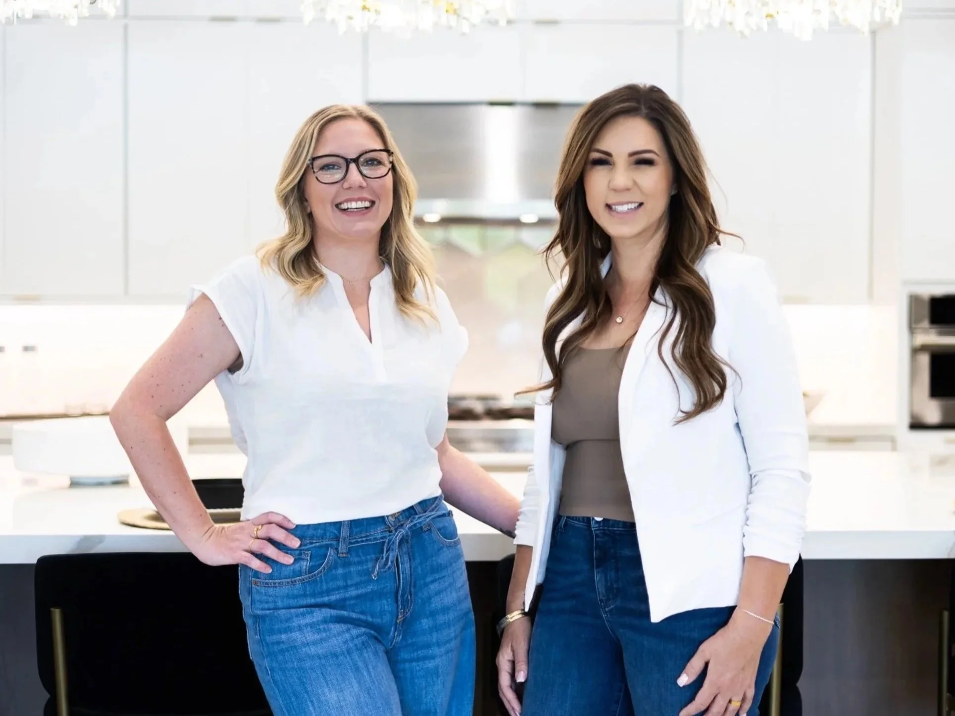 Two women standing in a bright kitchen, smiling and looking at the camera. One has blonde hair and glasses, the other has brown hair and is holding a tablet.