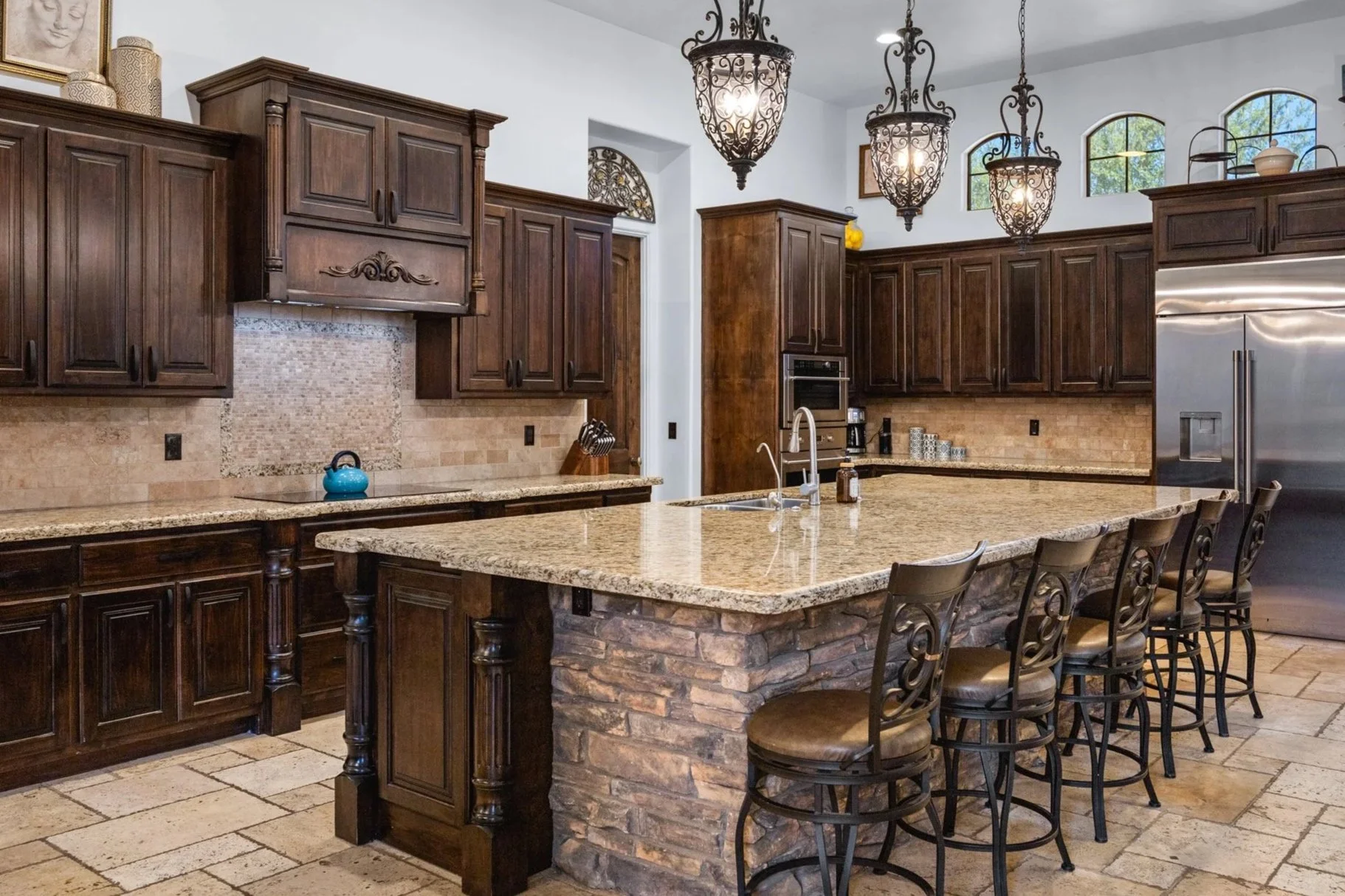 A spacious kitchen with dark wooden cabinets, a large granite island with a stone base, metal bar stools with cushioned seats, pendant chandeliers hanging from the ceiling, a stainless steel refrigerator, and a granite backsplash.