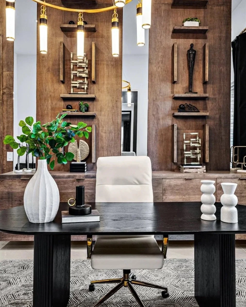 Modern office with a large dark wood desk, a white chair, and decorative items including a vase with green leaves, white vases, and artwork on wood-paneled walls with shelves and hanging lights.