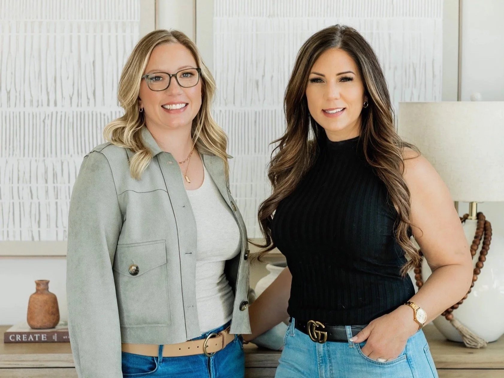 Two women standing indoors, smiling at the camera. One has blonde hair, glasses, and is wearing a light green blazer over a white top. The other has long dark hair, and is wearing a black sleeveless top and light blue jeans with a black Gucci belt. There is a white wall behind them, a bedside lamp, books, and decorative objects.