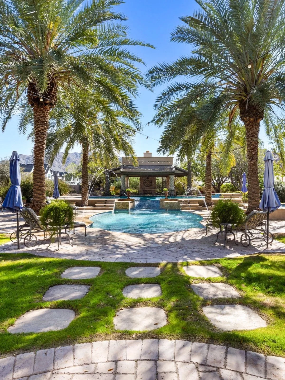 A resort pool area with two tall palm trees flanking the scene. In the background, there is a small pavilion with water features, including fountains and waterfalls, and steps leading into the pool. Lounge chairs with blue umbrellas are arranged around the pool, and a stone pathway leads toward the area. The sky is clear and sunny.