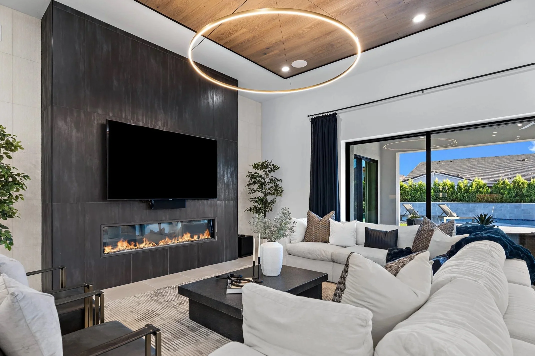 Modern living room with a black tiled wall featuring a fireplace and a flat-screen TV above it. There is a unique circular LED light fixture on the wooden ceiling. The room has large sliding glass doors opening to an outdoor patio with lounge chairs and trees. The interior has white sofas, a black coffee table, and decorative pillows.