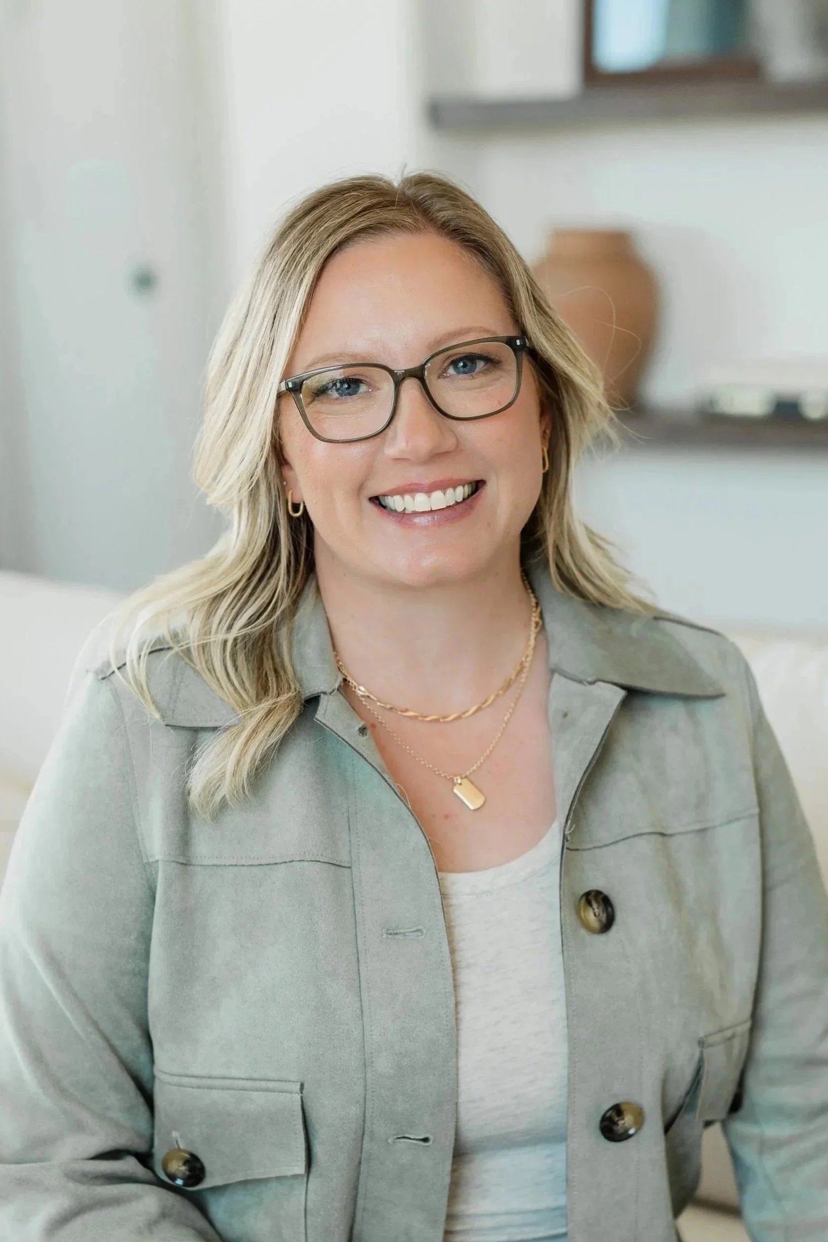 A smiling woman with blonde hair, glasses, wearing gold jewelry, a light green jacket, and a cream-colored top in an indoor setting.
