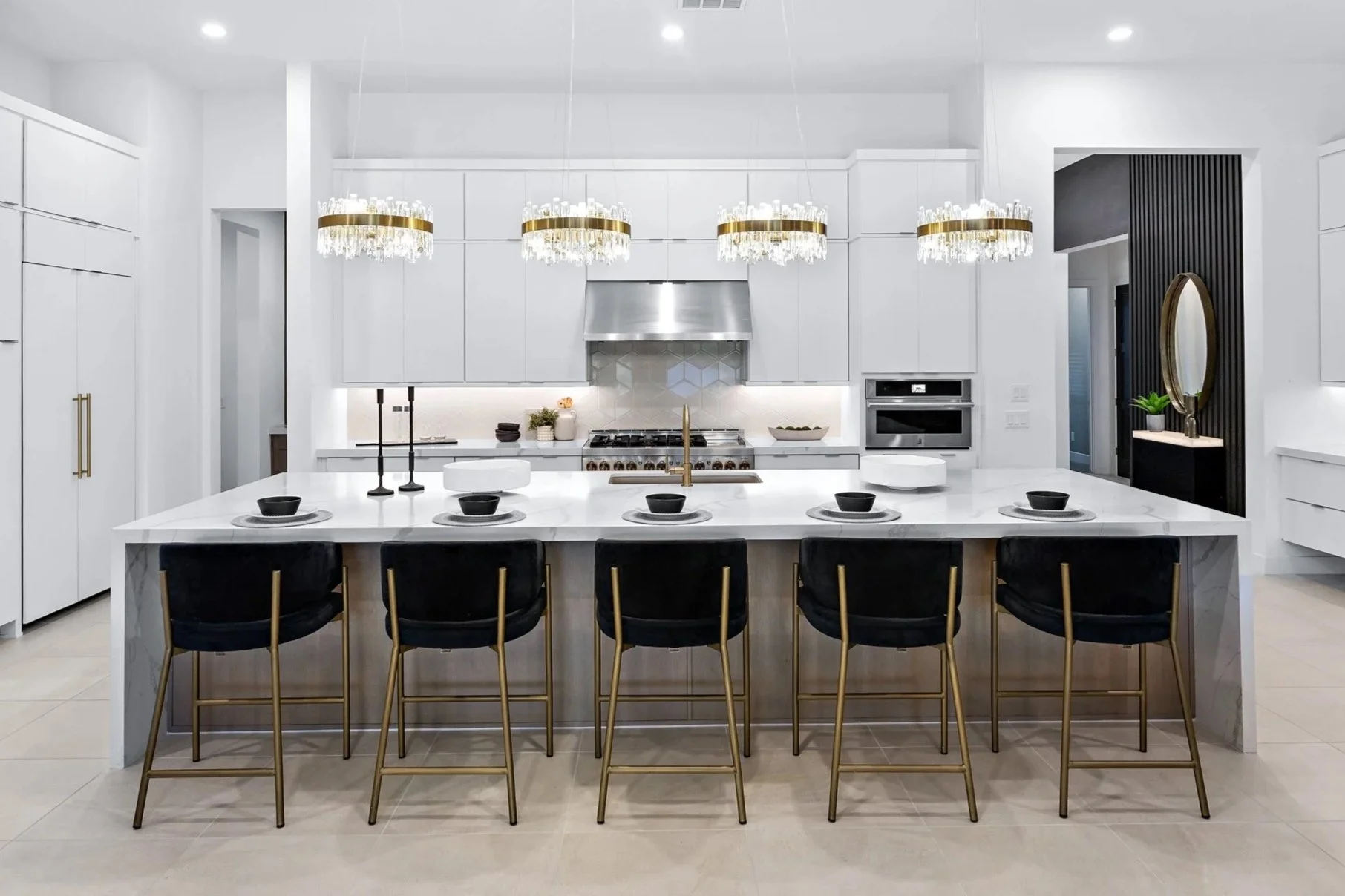 Modern white kitchen with marble island, black chairs, chandelier lighting, and stainless steel appliances.