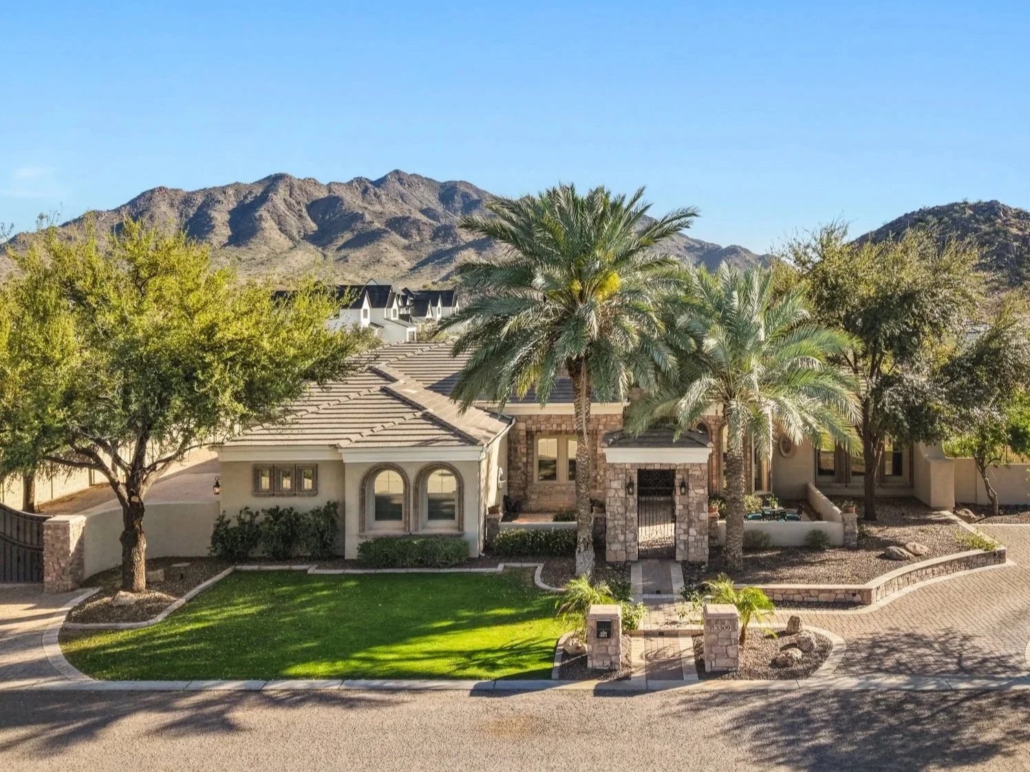 A luxurious house with stone and stucco exterior, surrounded by palm trees and greenery, with mountains in the background.