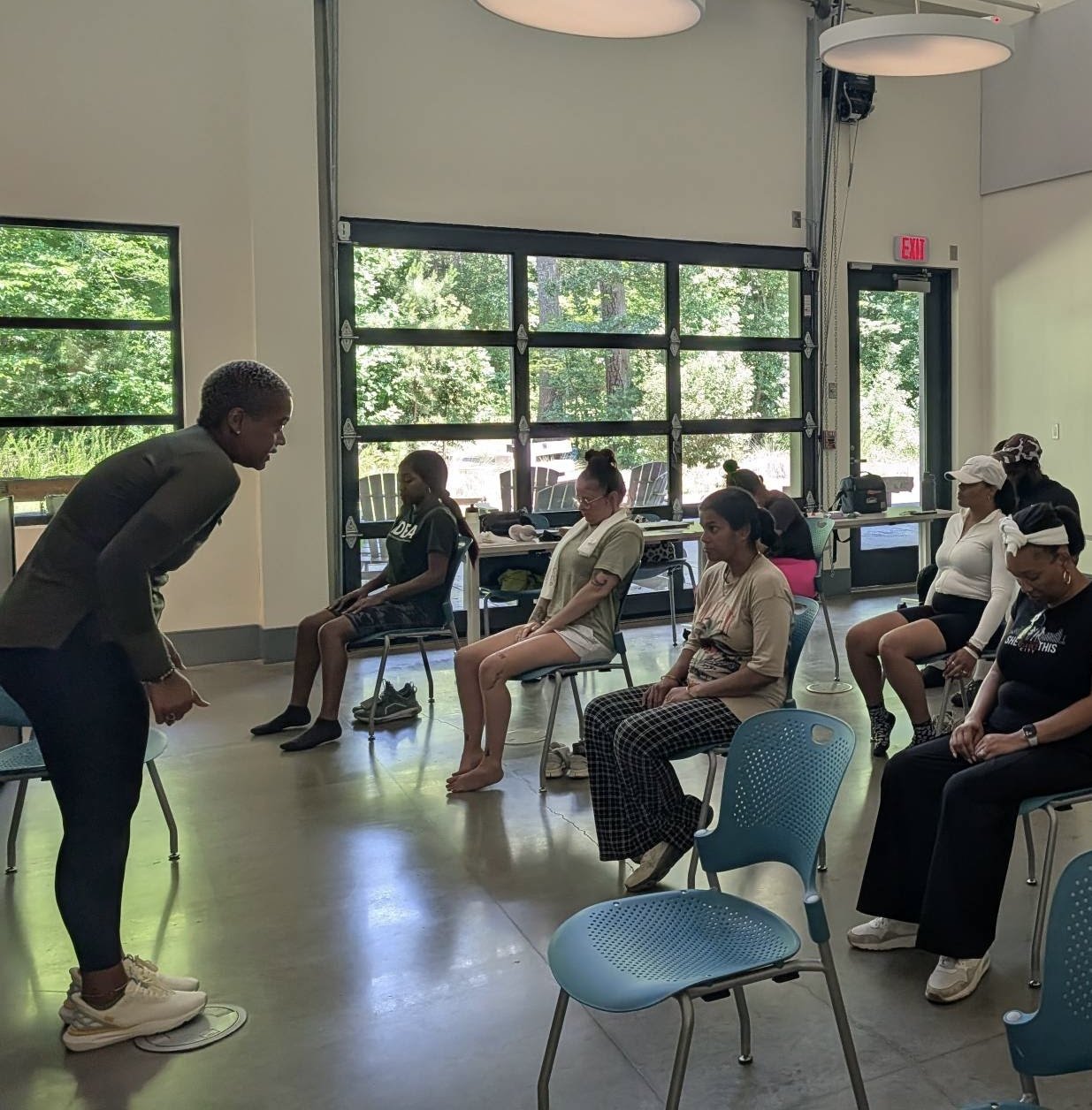 A group of women sitting on chairs in a room with large windows, a man and a girl sitting in front of them, and a woman standing near the man and girl, all engaged in an activity.