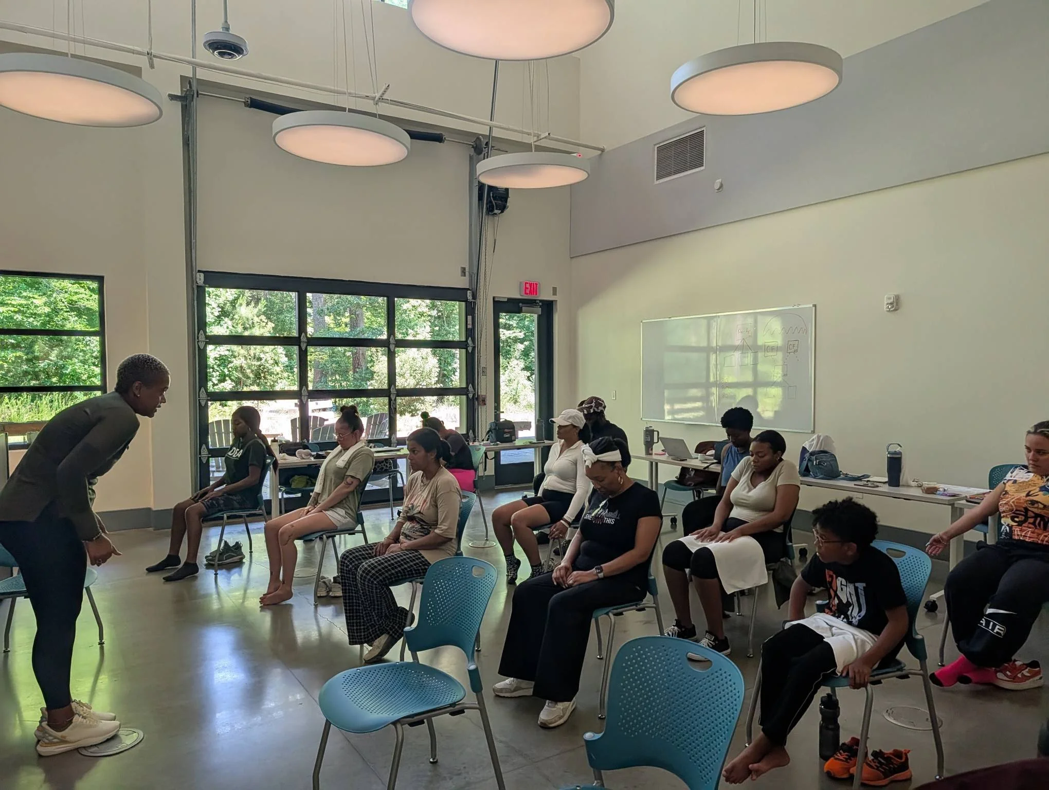 A group of people sitting in chairs in a bright room with large windows, white walls, and hanging circular lights, participating in a meditation or relaxation session with some individuals with their eyes closed.