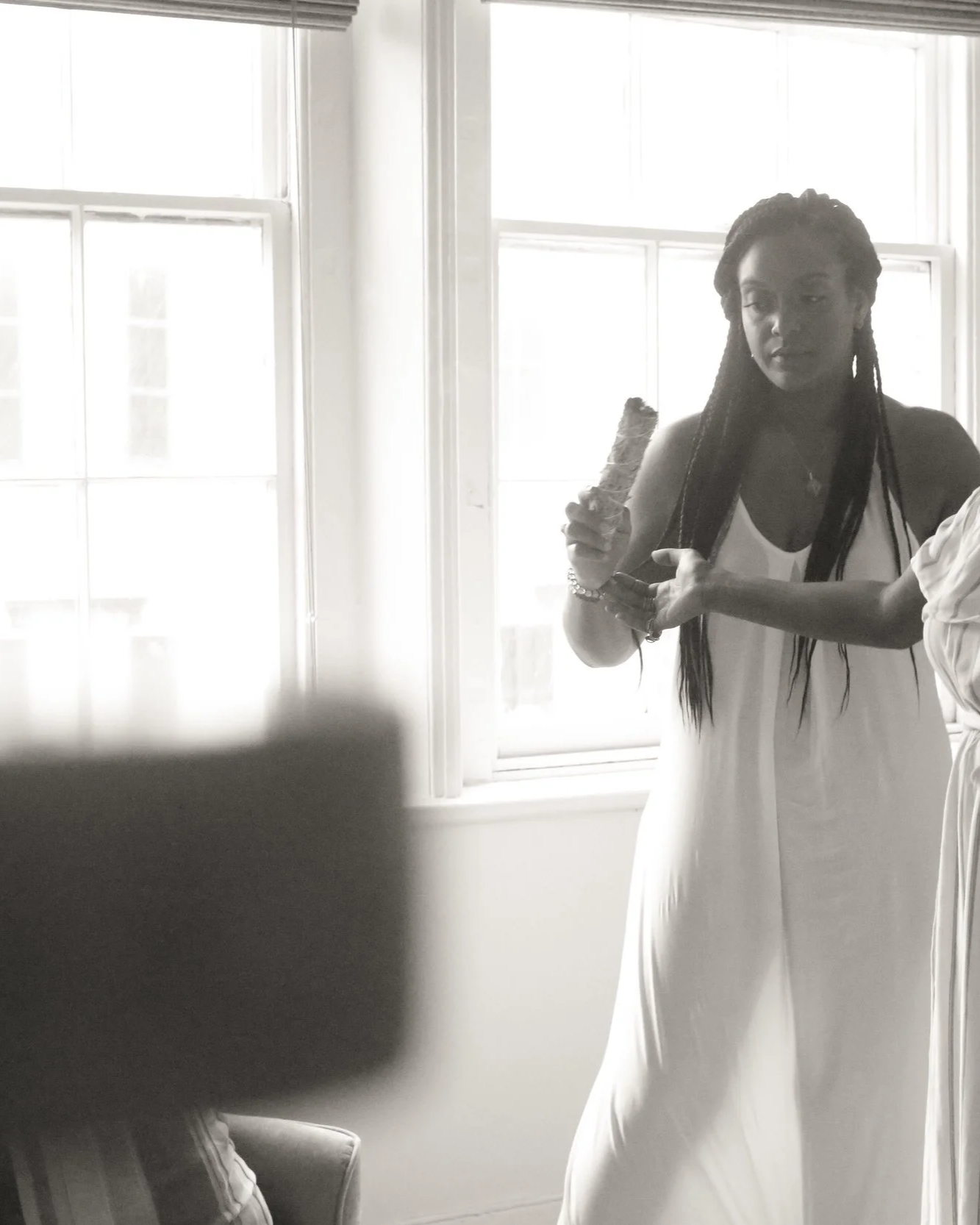 A woman wearing a white dress is standing near a window, holding a piece of cake, with sunlight coming through the window illuminating the scene.