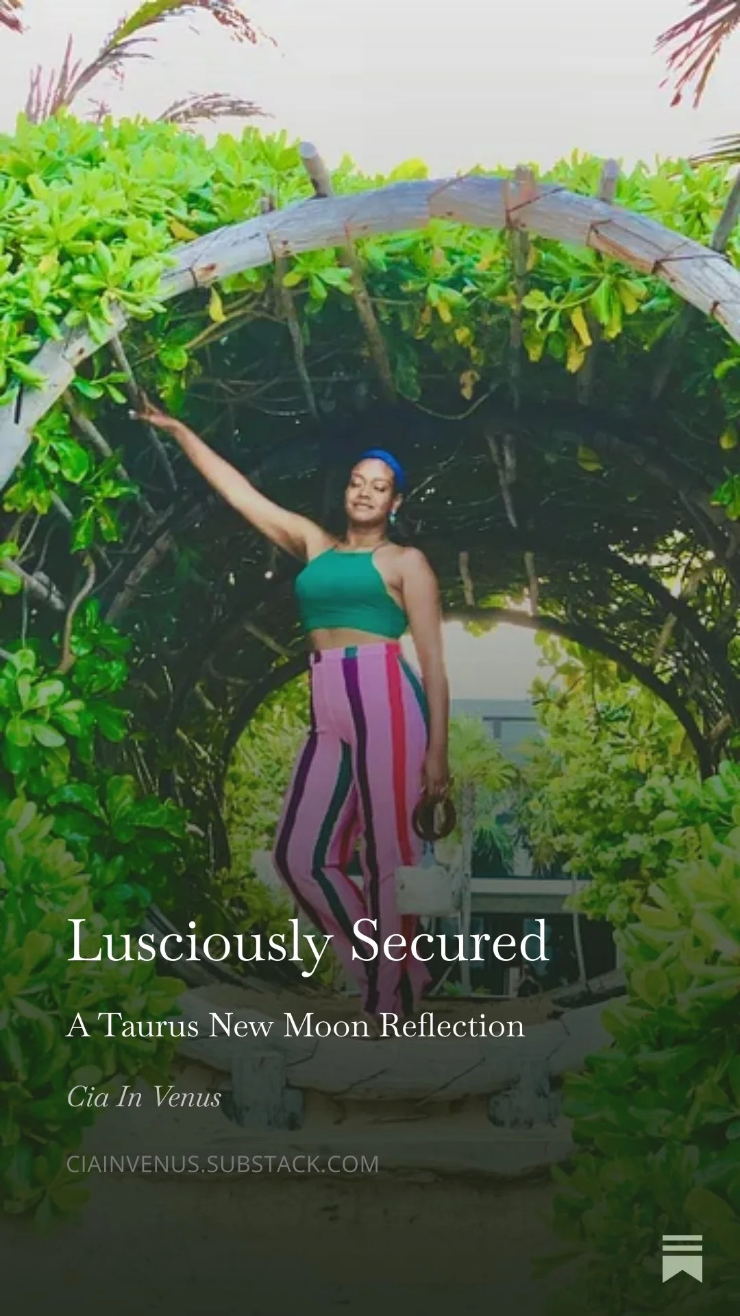 Woman standing under a vine-covered archway during daytime, wearing colorful striped pants and a green top, holding a small handbag.