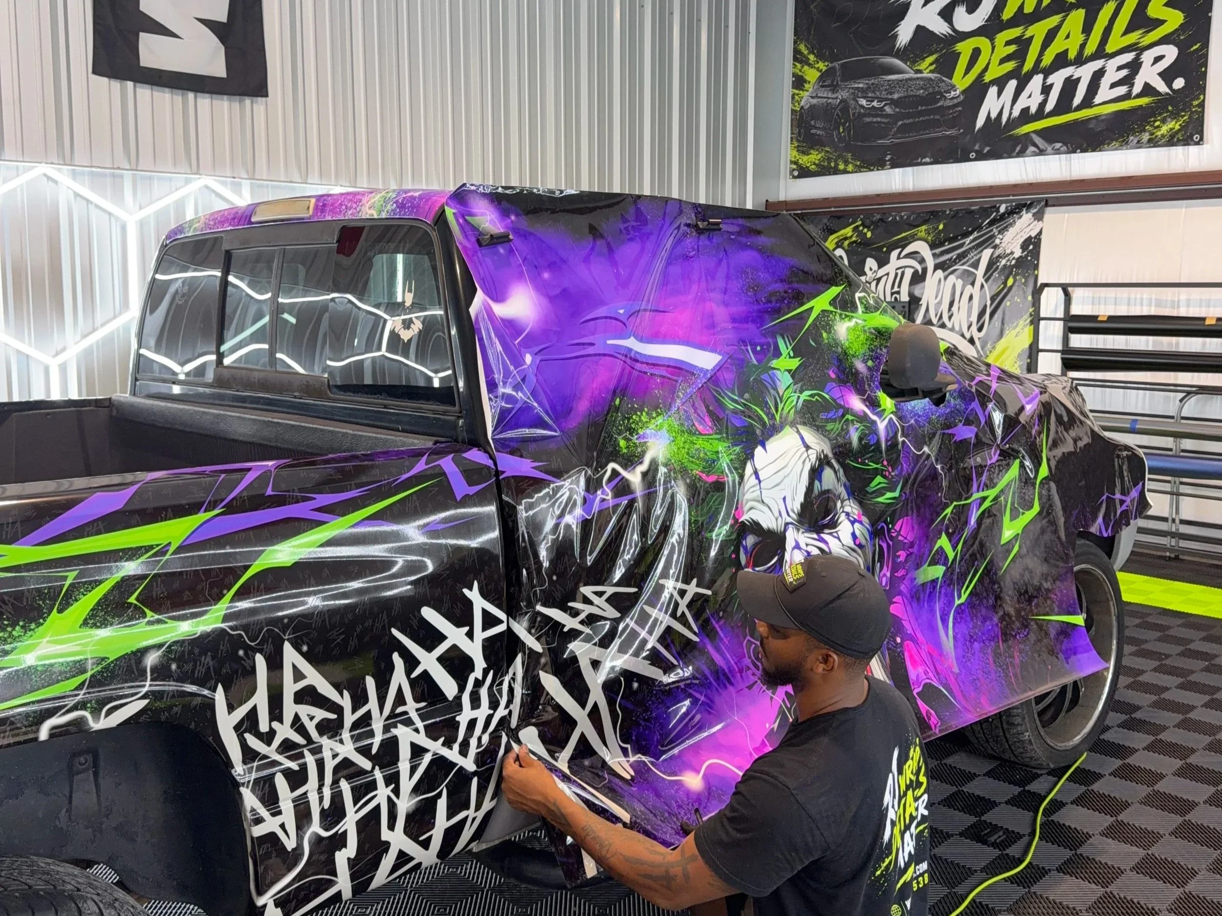 Person working on a colorful, graffiti-style vehicle wrap in an industrial setting, with banners promoting automotive detailing.