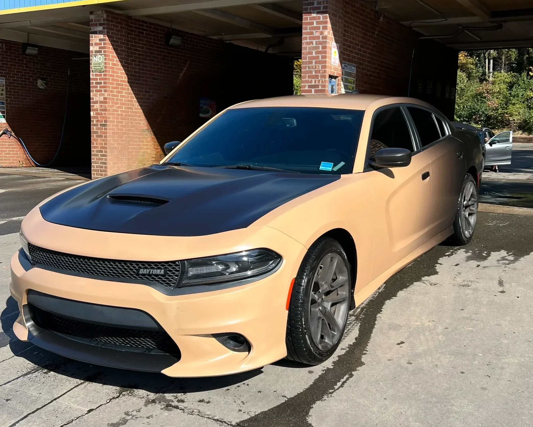 A beige and black Dodge Daytona car parked in a parking lot near a brick wall.