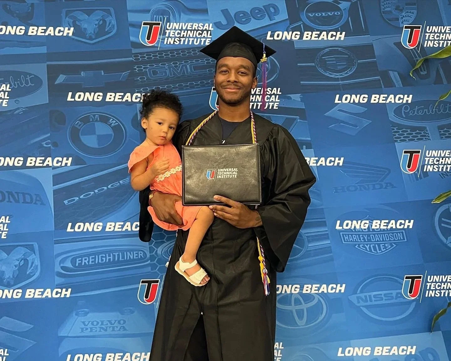 A young man in a graduation gown and cap, holding a diploma, standing next to a young girl in a pink dress. They are in front of a blue backdrop with logos and the words 'Universal Technical Institute' and 'Long Beach.'