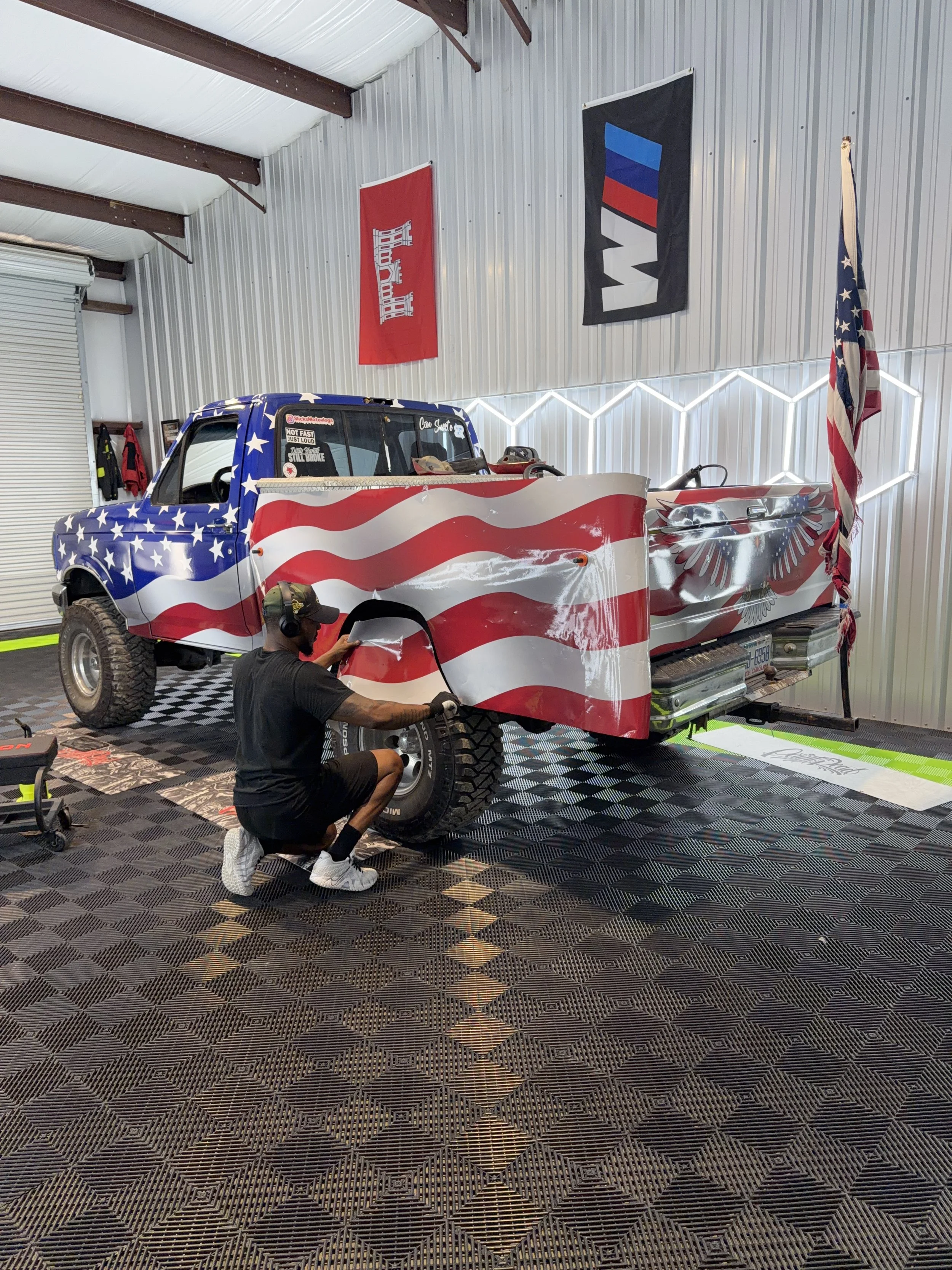 A person kneeling next to a truck with an American flag paint job inside a garage with patriotic flags hanging on the wall.