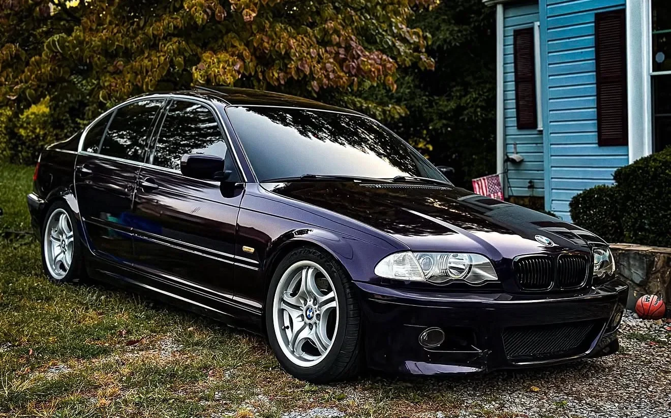 Black BMW sedan parked on grass in front of a blue house with brown shutters and a basketball