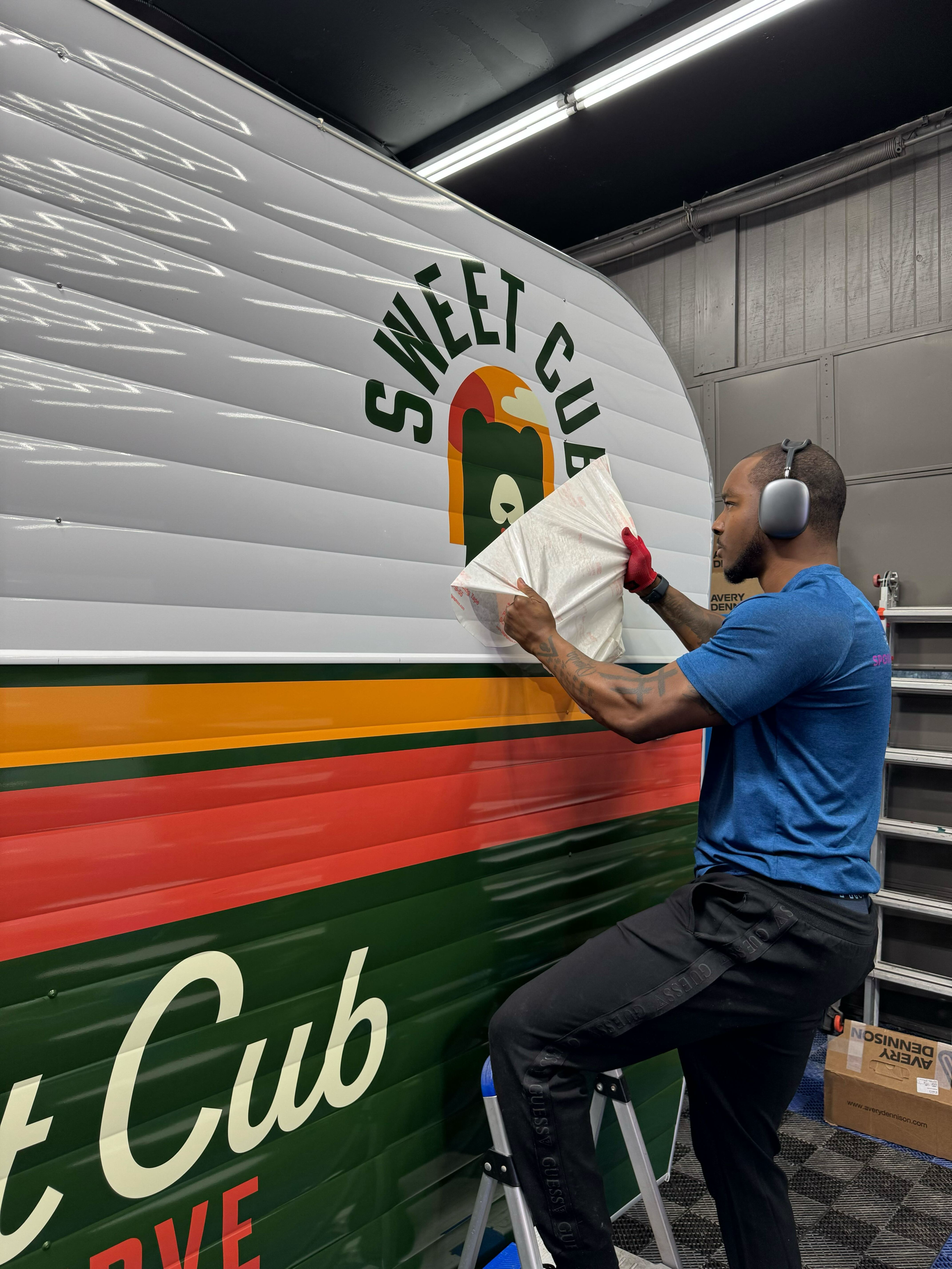 A man with a bald head and tattoos is standing on a step stool, applying a large vinyl decal to the side of a truck. He is wearing a blue shirt, black pants, and gray earmuffs for protection.