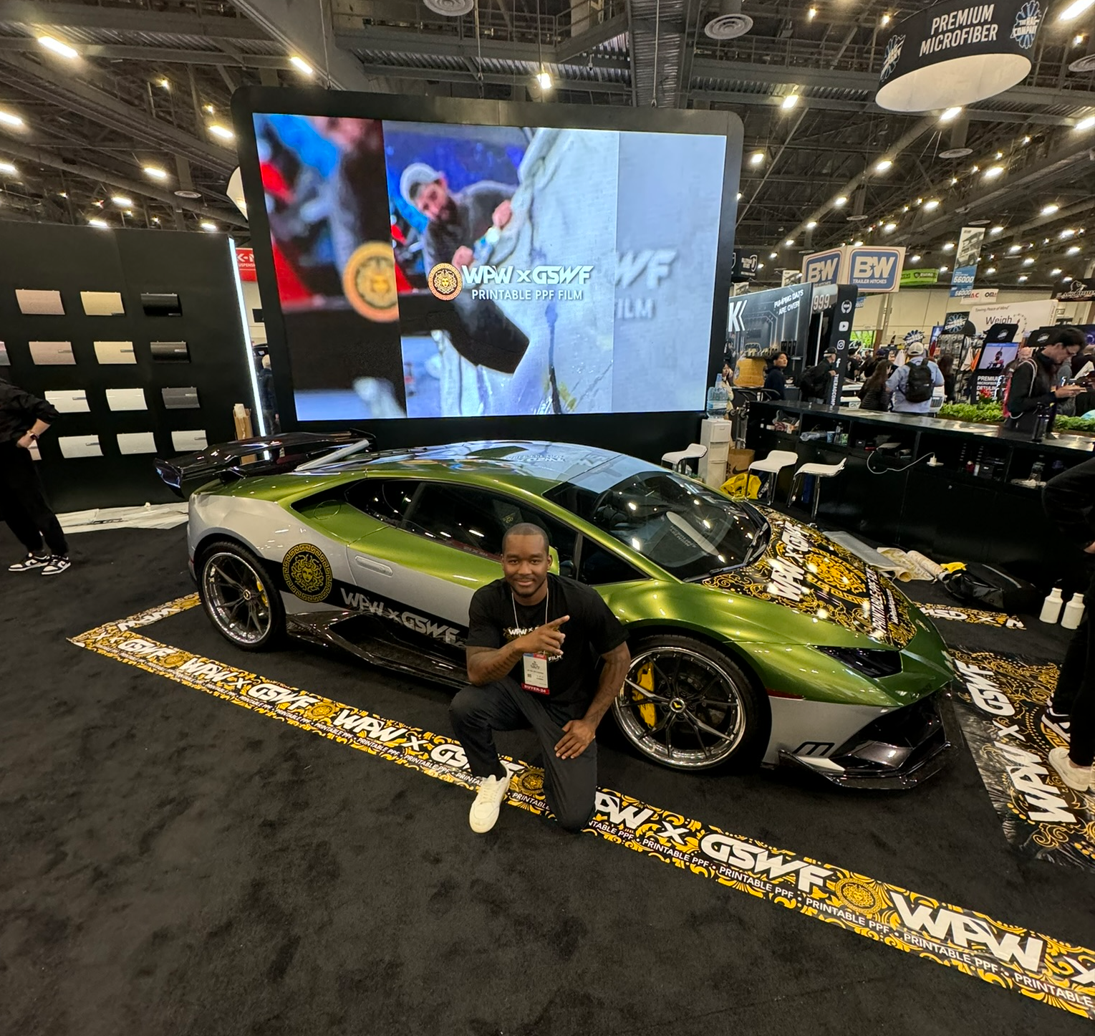 A man kneeling and smiling next to a green and black Lamborghini sports car at an indoor exhibition. The car is on a printed mat with logos and text, in front of a large display screen showing a person snowboarding and the text 'WPW x GSWF Printable PPF Film.' The background shows other booths and visitors at the event.