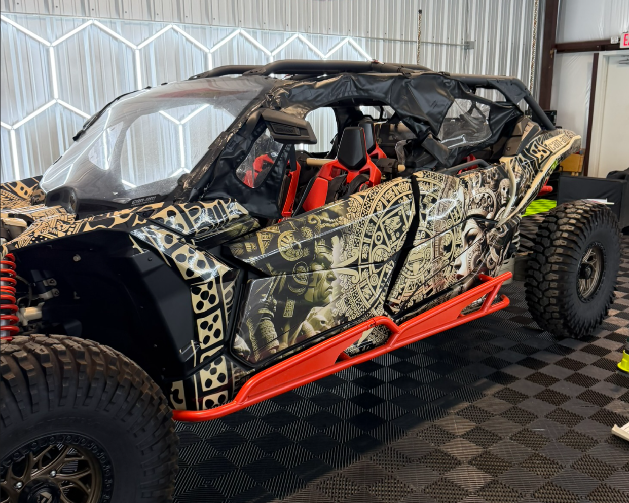 Off-road race car with black and gold Aztec-style artwork and red accents, inside a garage.