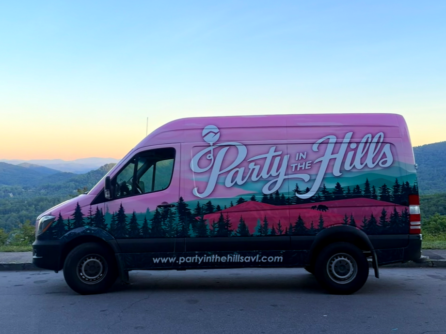 Purple decoration van with trees and hills design and text 'Party in the Hills' parked on roadside with mountains in the background at sunset.