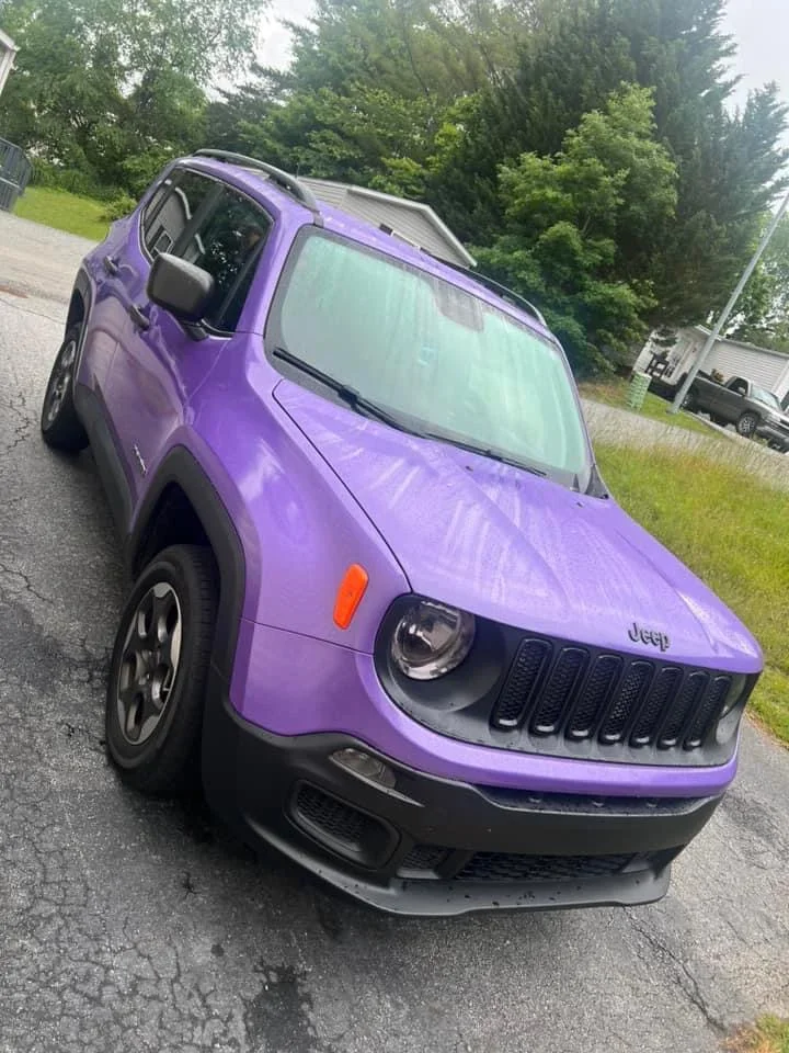 A purple Jeep Renegade parked on a cracked asphalt driveway with green trees and a house in the background.
